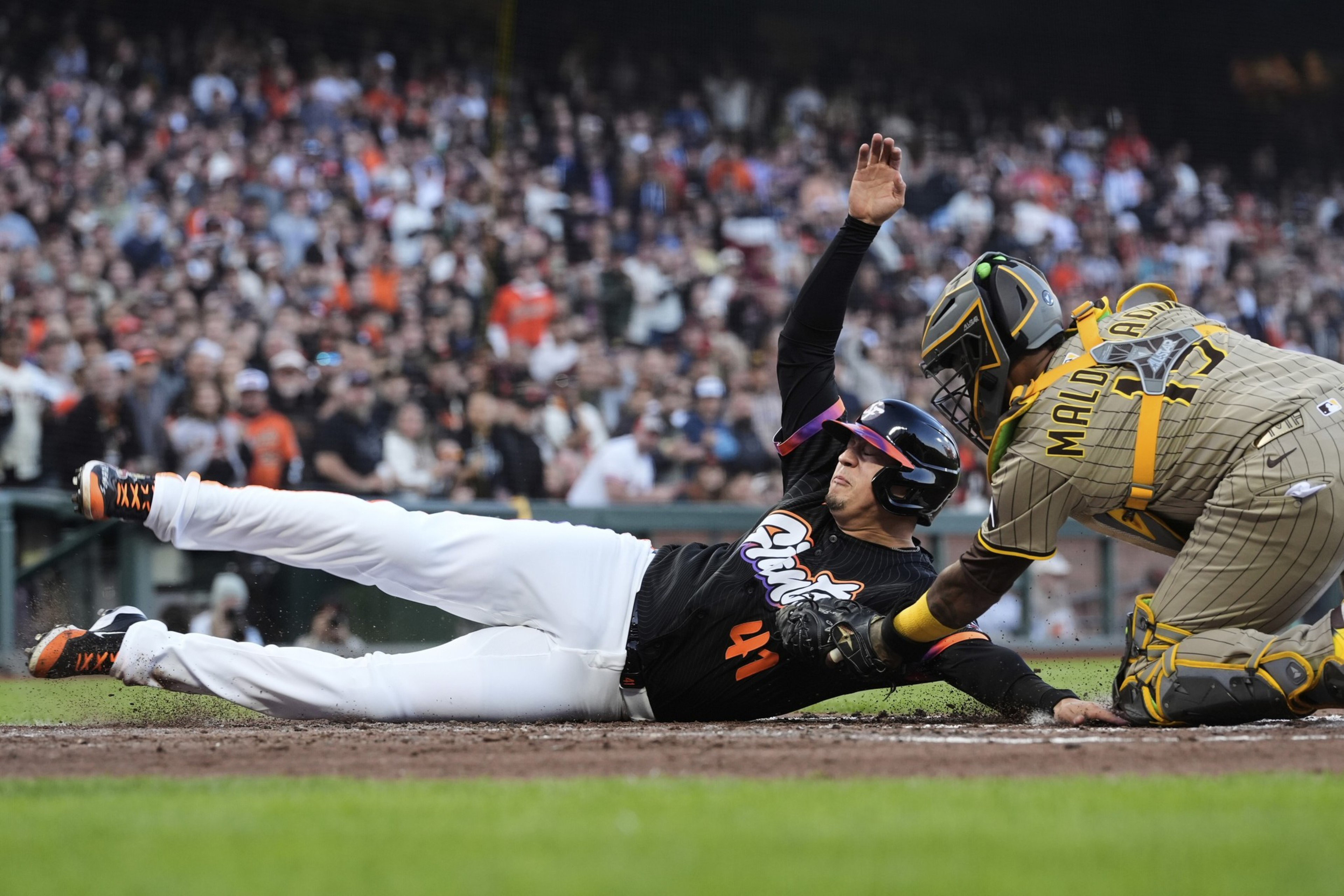 A baseball player in black slides into home plate as a catcher in tan gear reaches to tag him, with a large crowd watching in the background.
