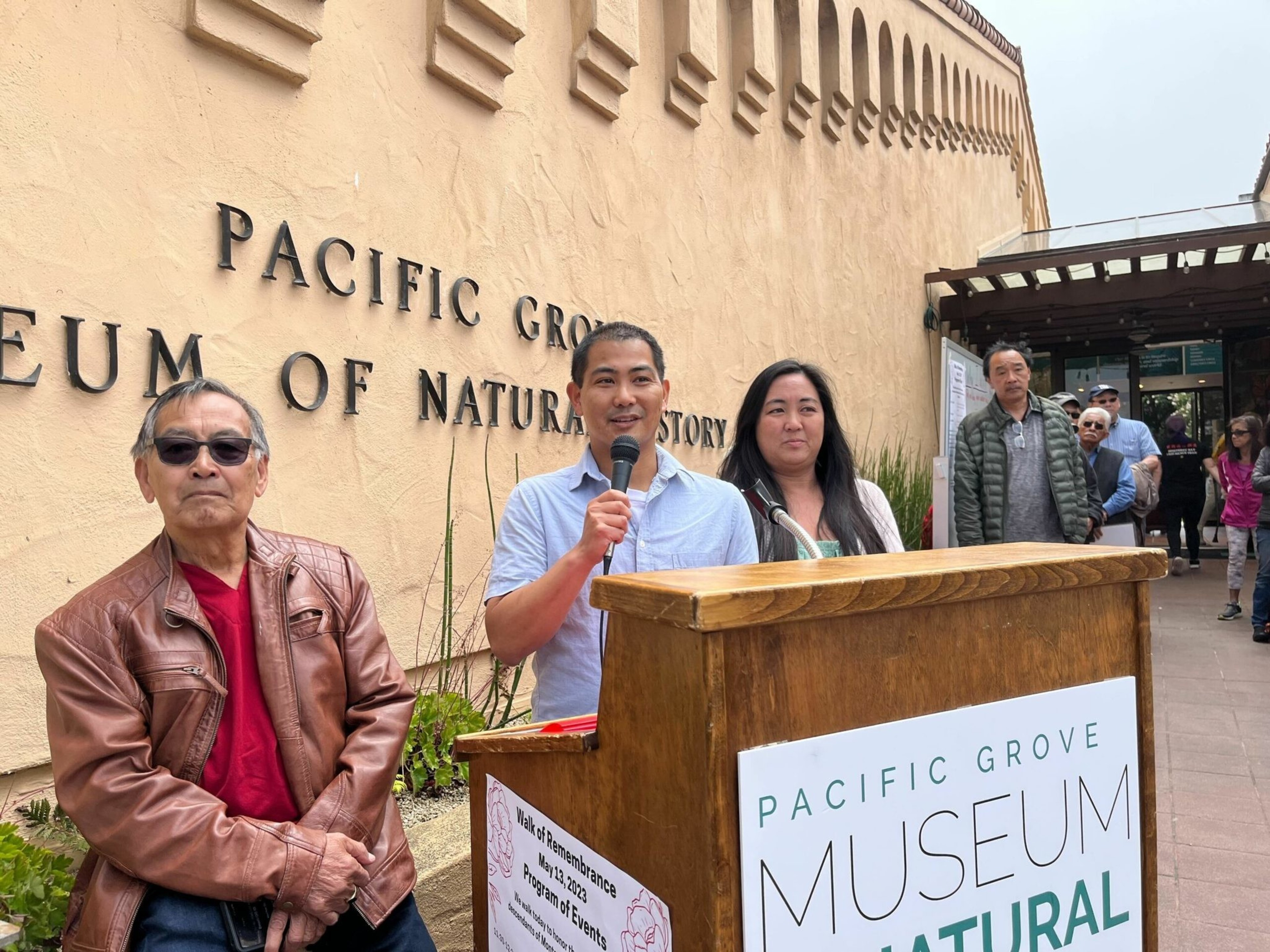 A man speaks into a microphone at a wooden podium outside the Pacific Grove Museum of Natural History, with three people standing nearby.