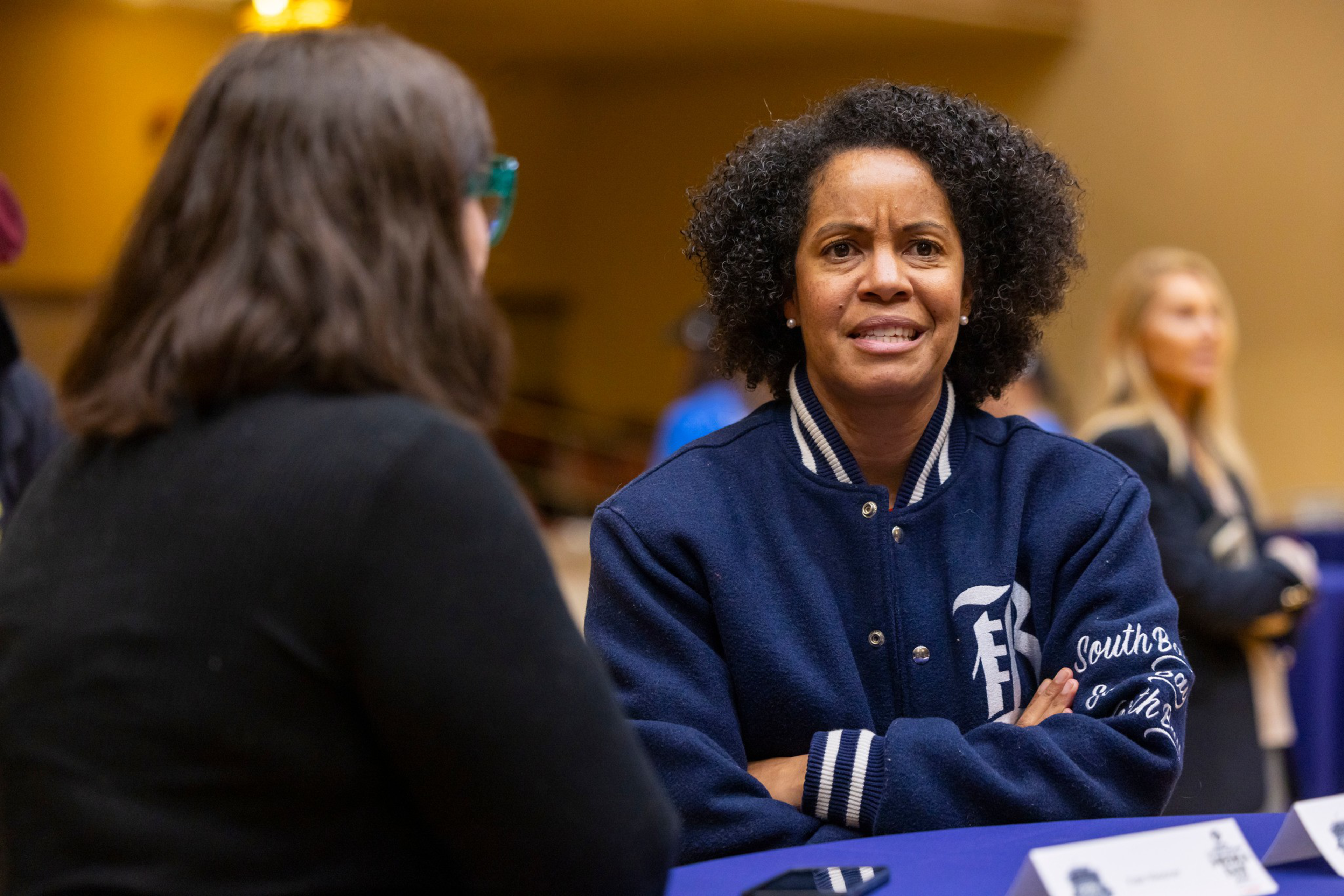 A woman with curly hair and a blue jacket speaks with another woman with long dark hair across a table in a busy indoor setting.