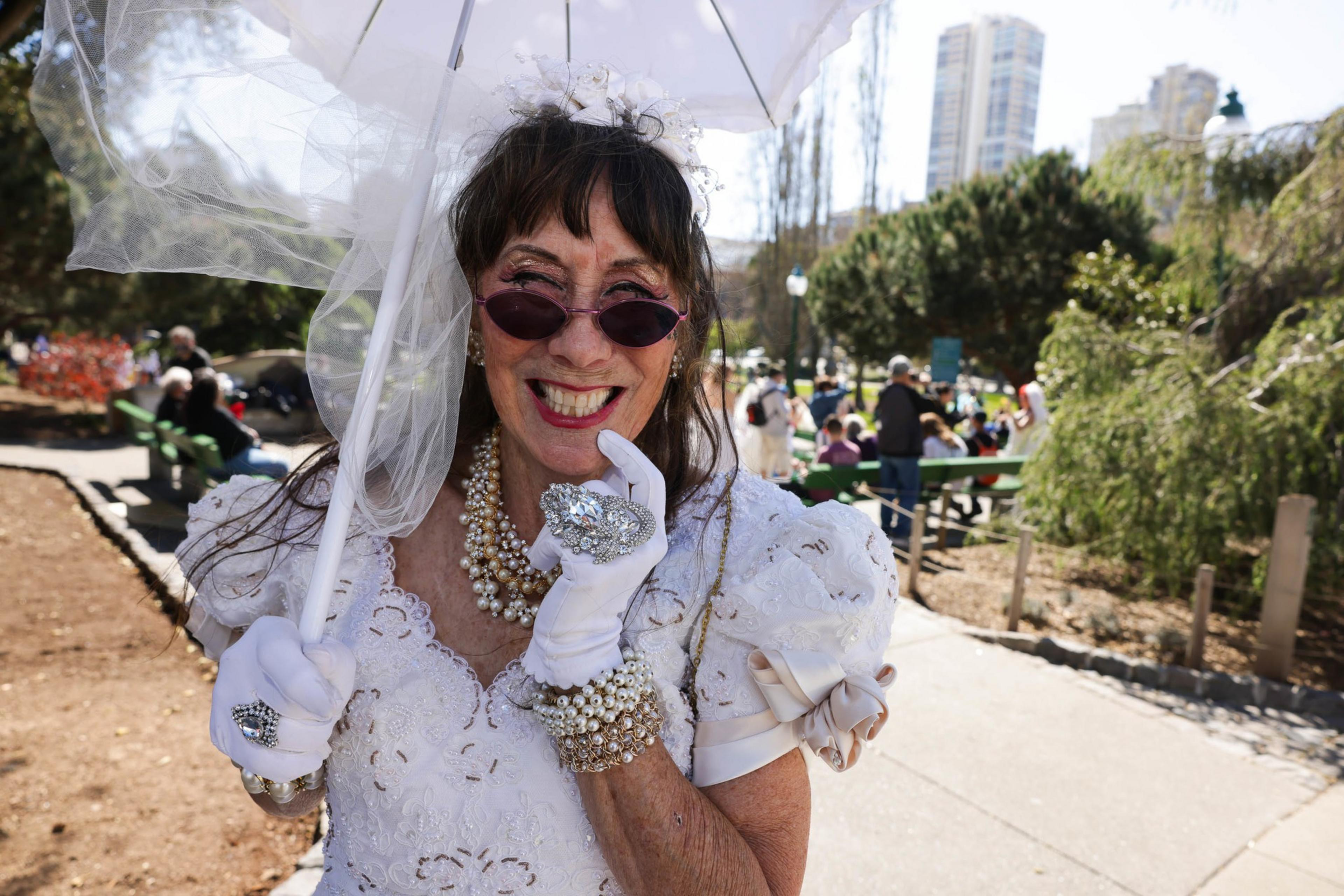 A smiling woman in a white bridal outfit wears sunglasses and pearls and holds an umbrella in an outdoor setting with people in the background.