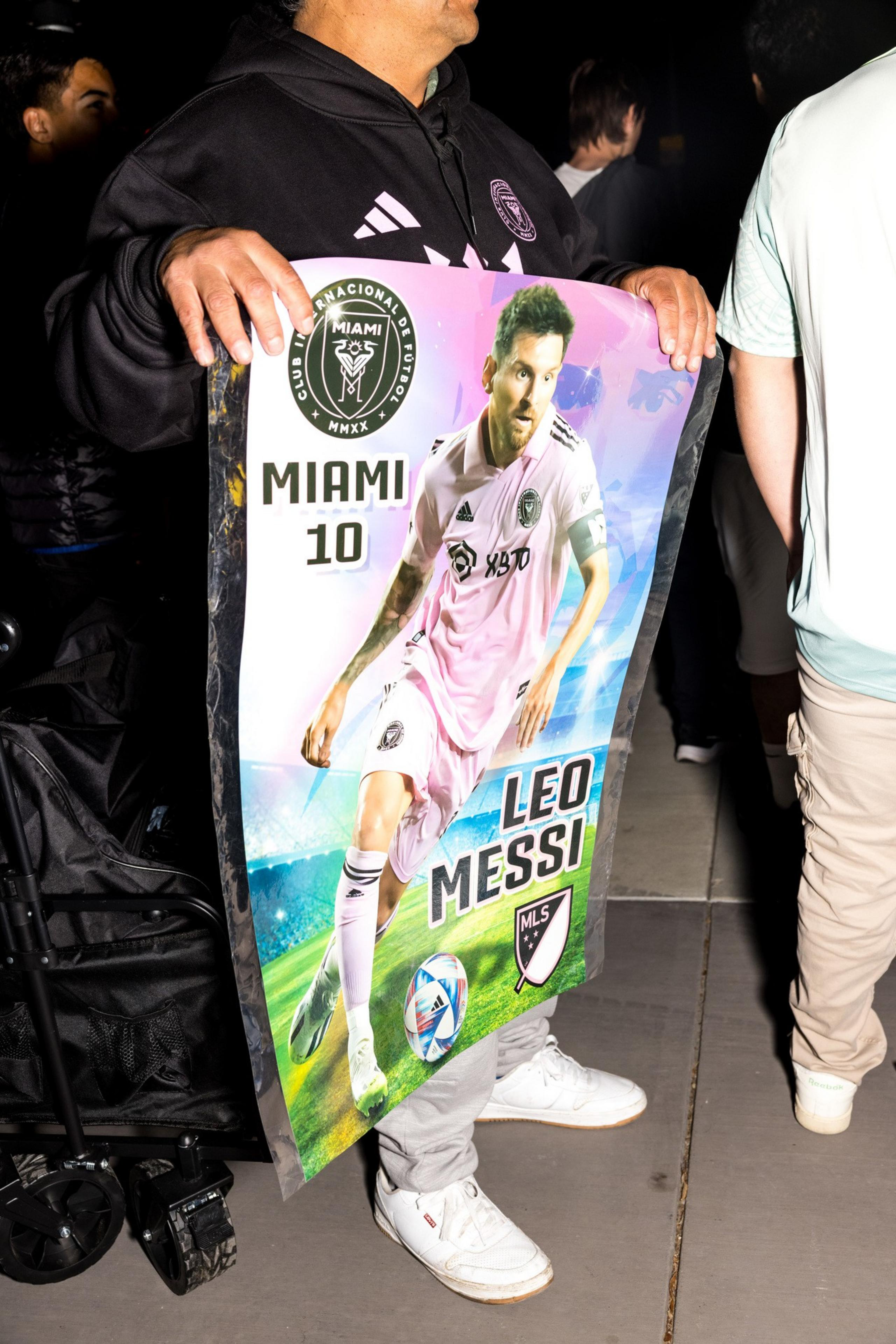 A person holds a Miami soccer poster featuring player number 10, Leo Messi, in a pink uniform. The poster includes the MLS logo.