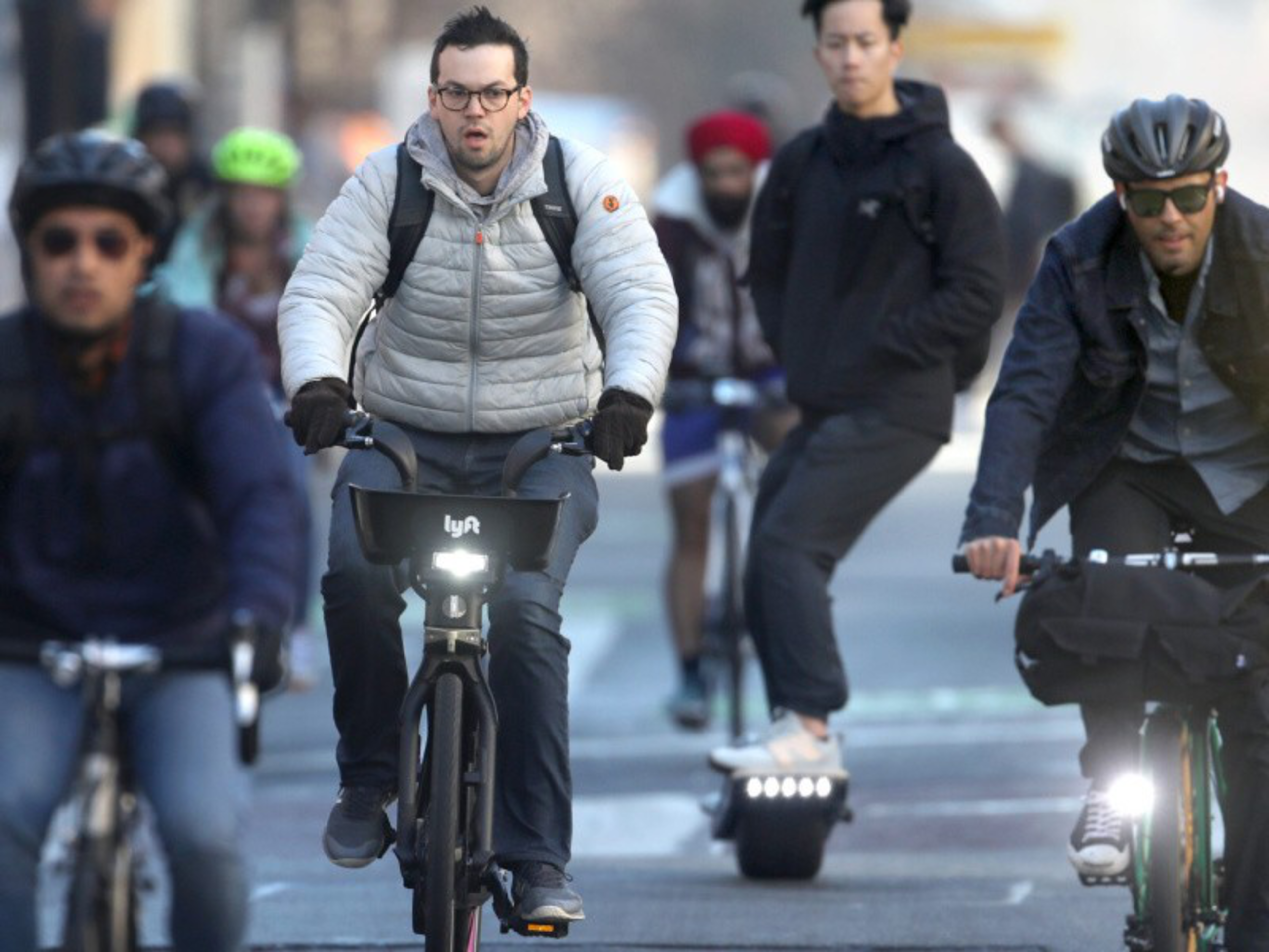 Several people are commuting on a city street, mostly on bicycles, with one person riding an electric one-wheeled scooter among them.