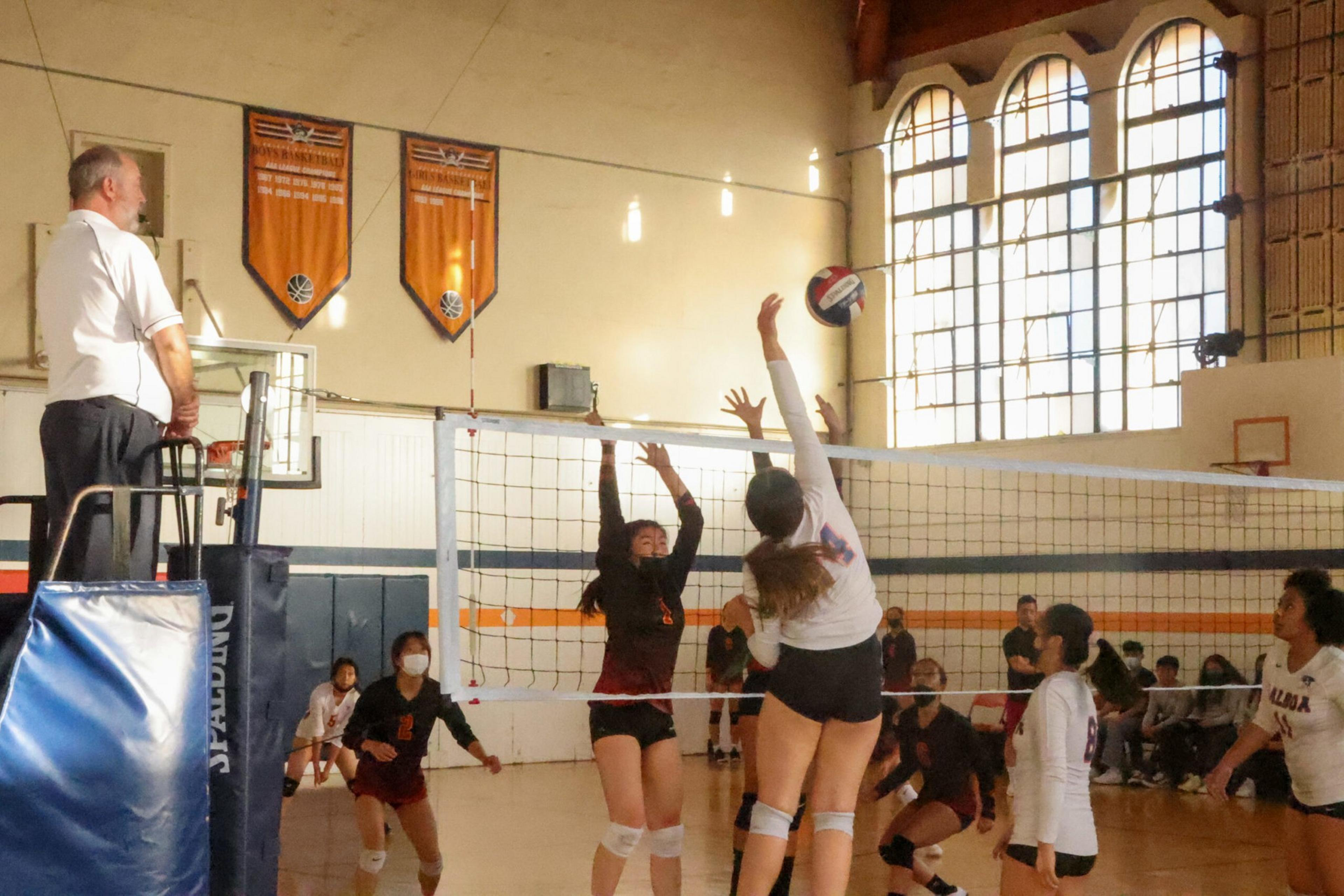 Players jump near the net, one hitting the volleyball while others attempt to block, with a referee watching from an elevated stand and spectators in the background.