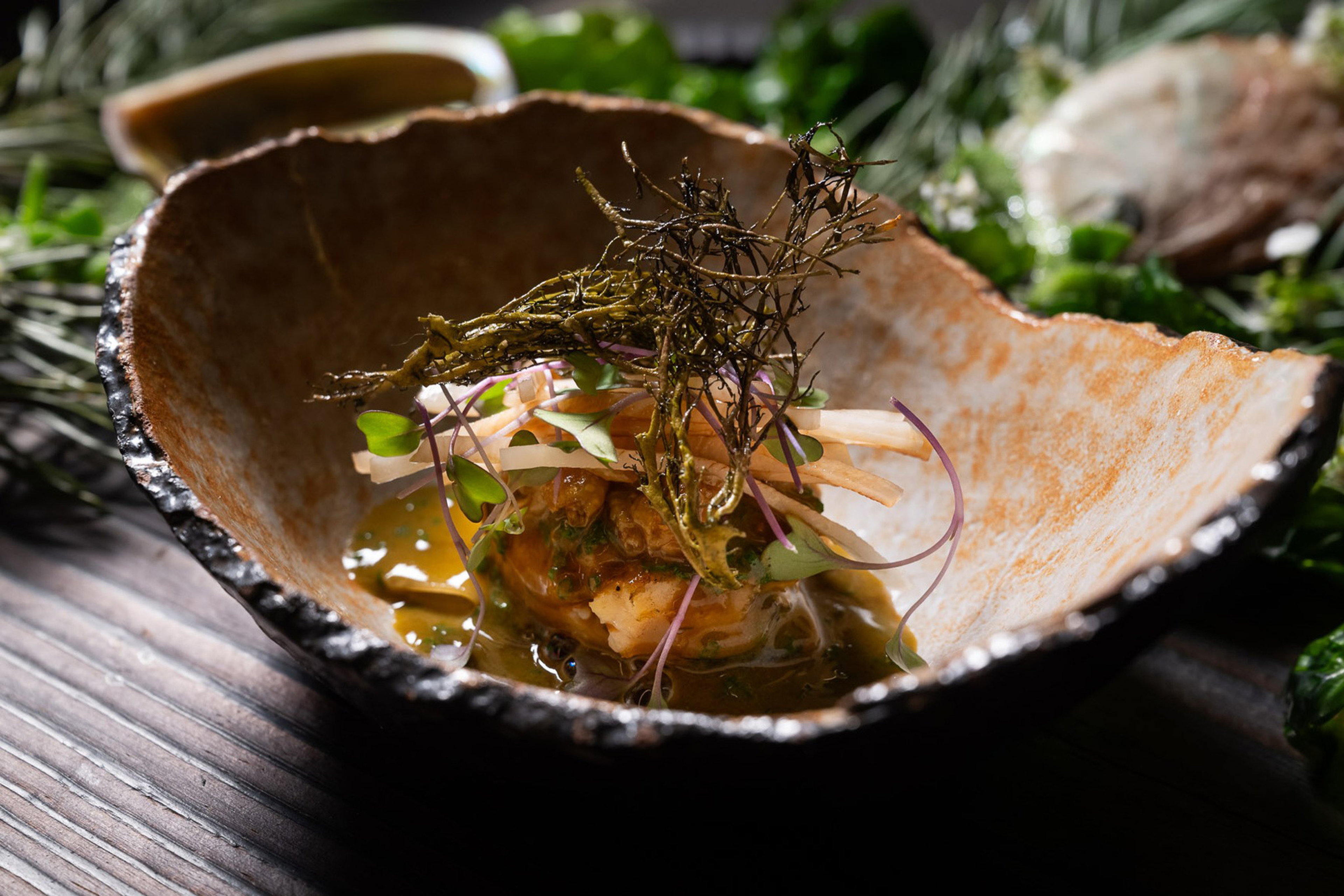 A rustic bowl contains a gourmet dish with greens, thinly sliced vegetables, and a sauce. The presentation is artistic, with a garnish of dried twigs.