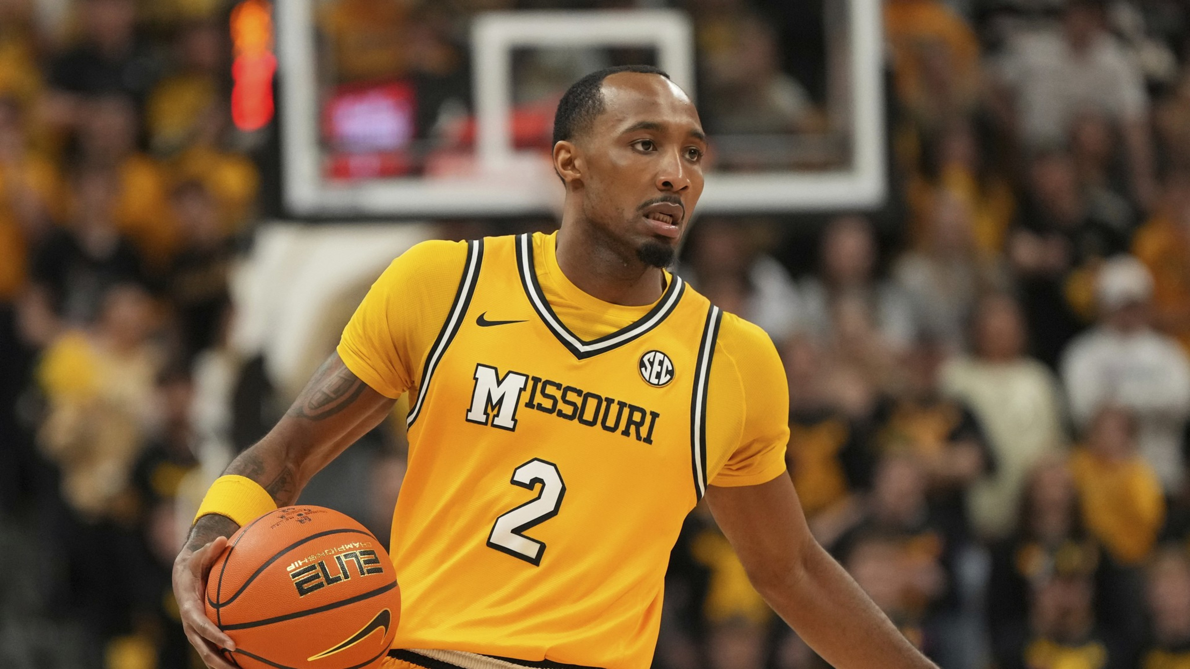 A basketball player in a yellow &quot;Missouri&quot; jersey with the number 2 is dribbling the ball on a court, amidst a blurred crowd background.
