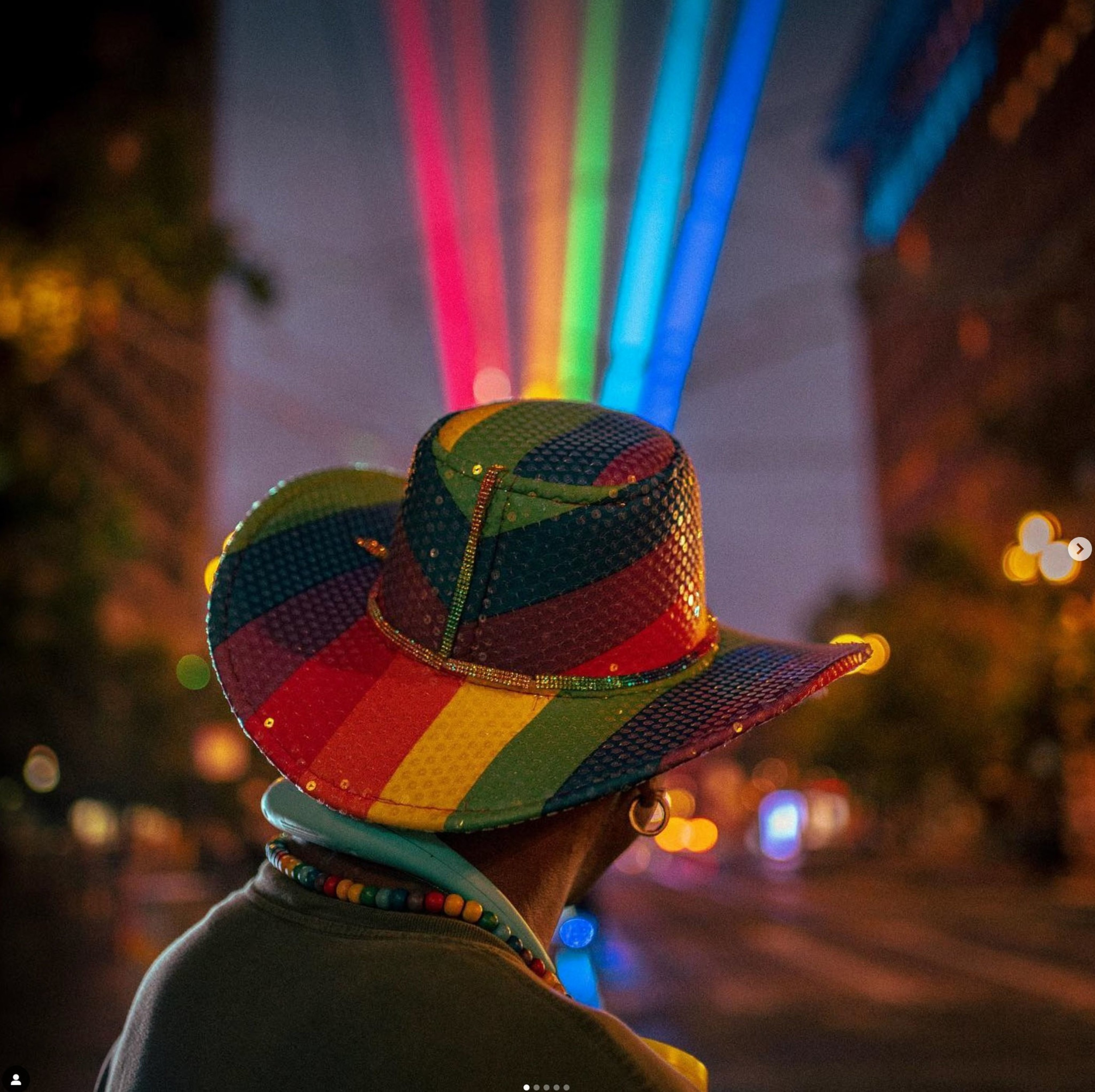 A person in a sequined, rainbow-striped hat stands on a city street, facing vibrant beams of colorful lights stretching across the night sky.