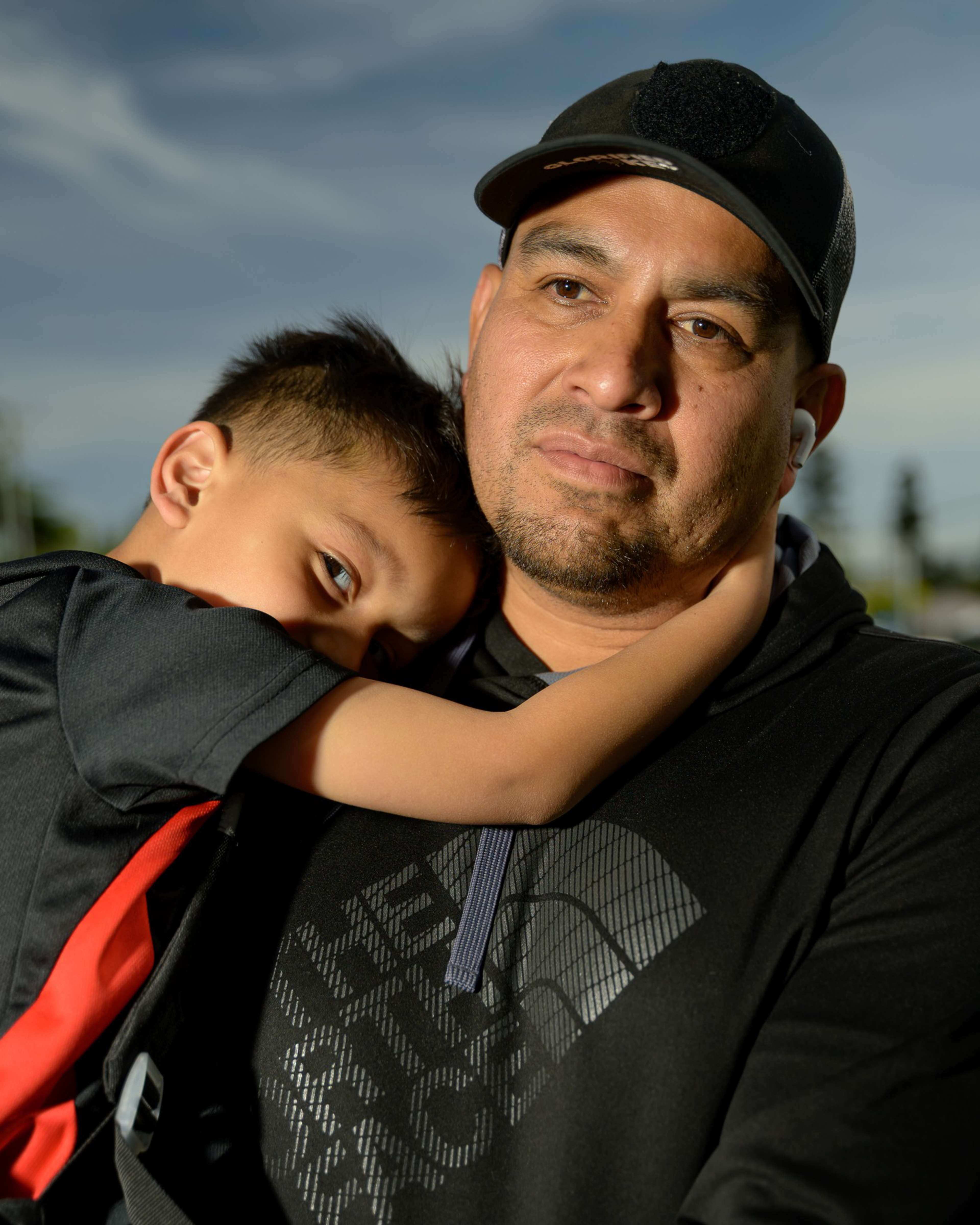 A man wearing a cap gently holds a young boy resting his head on the man's chest. They both have a calm expression, set against a sky background.