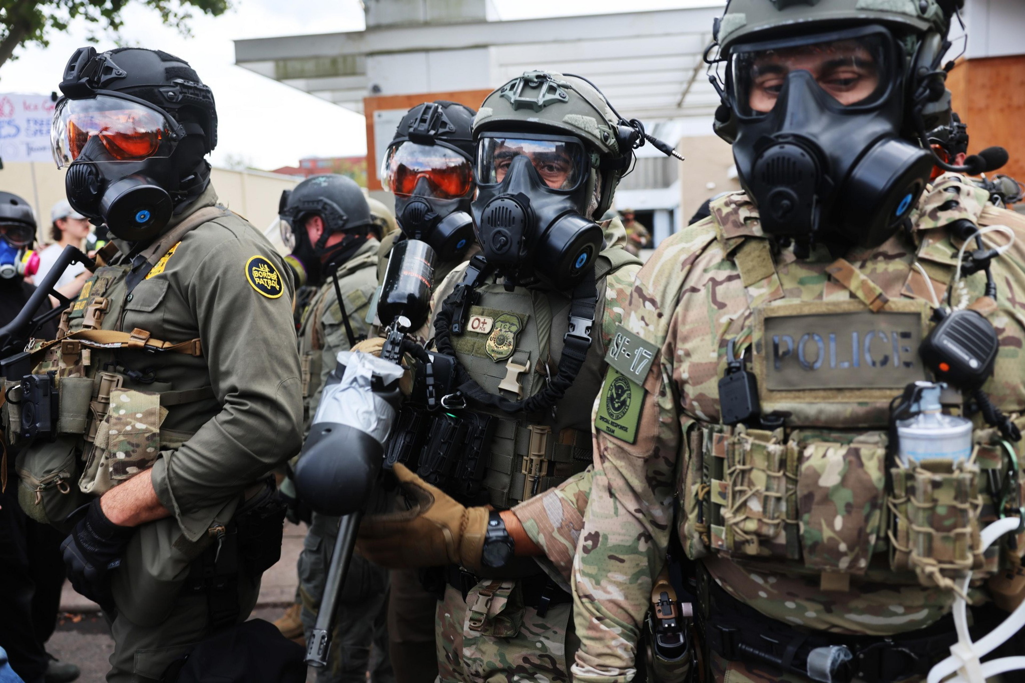 Four heavily armored police officers wearing tactical gear and gas masks stand closely together, appearing prepared for a crowd or protest.