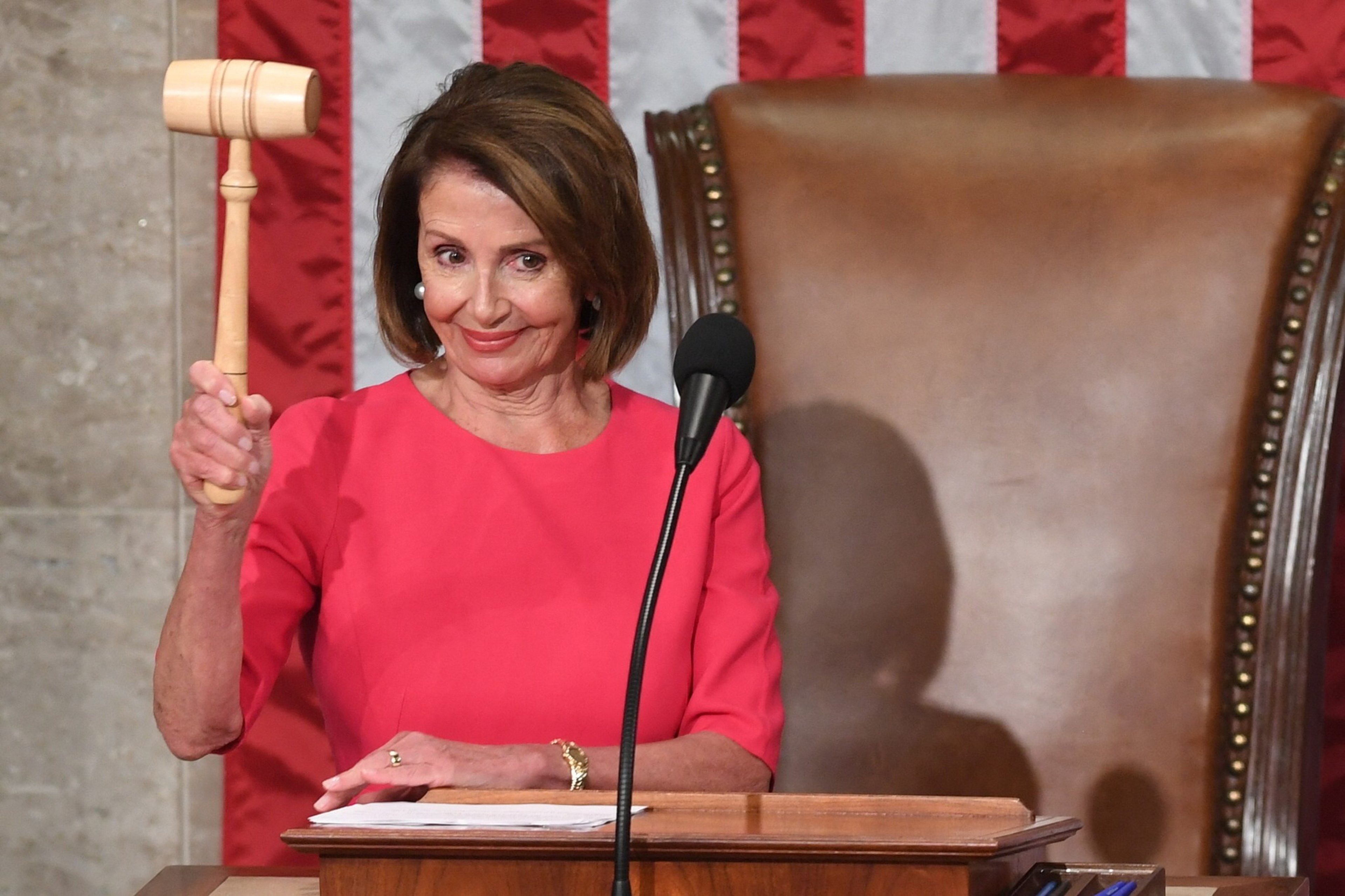 A woman in a pink dress holds up a wooden gavel, standing behind a podium with a microphone, with a large chair and American flag in the background.