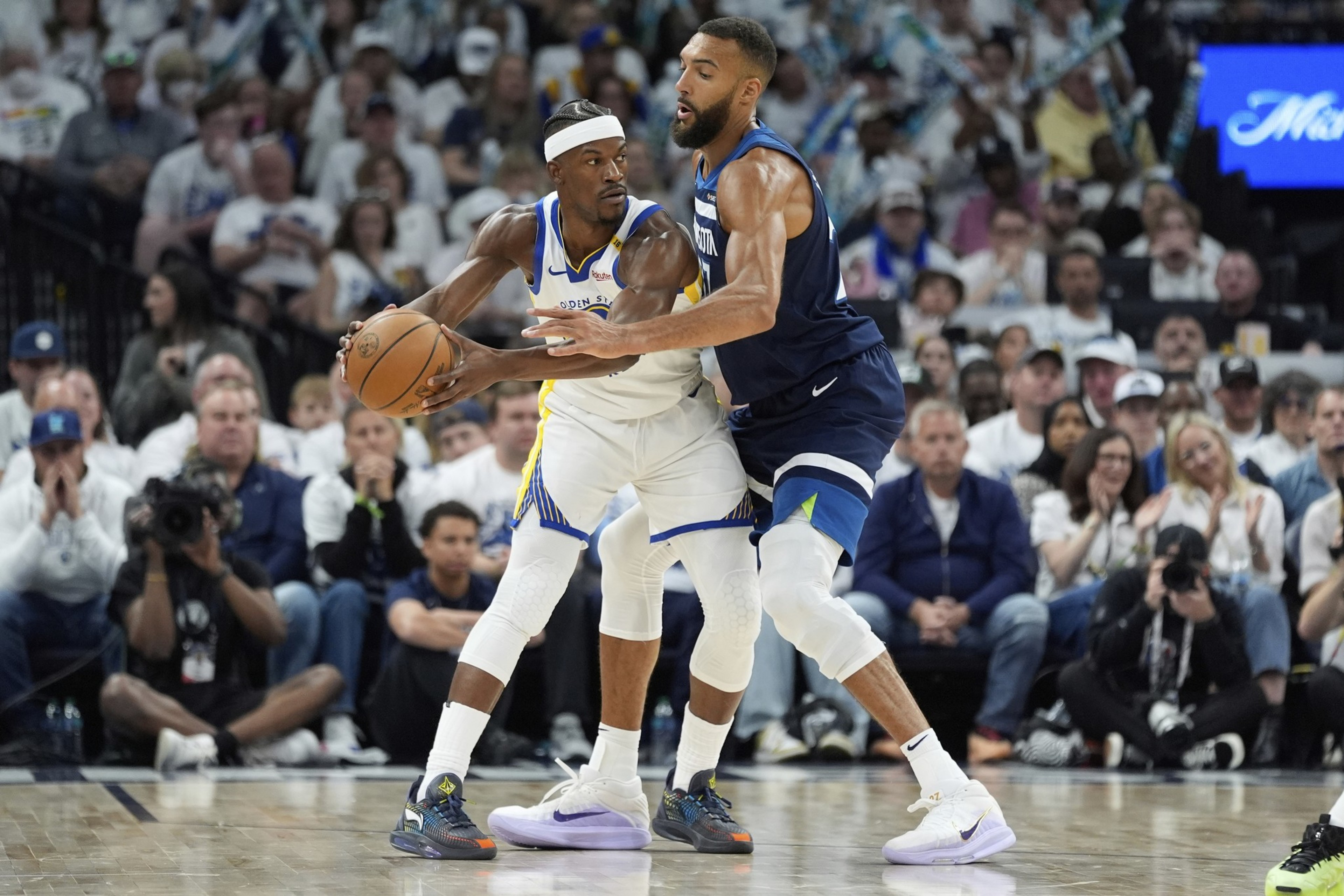 Two basketball players are on a court, one in a white jersey holding the ball, and the other in a dark jersey defending. A crowd watches intently.