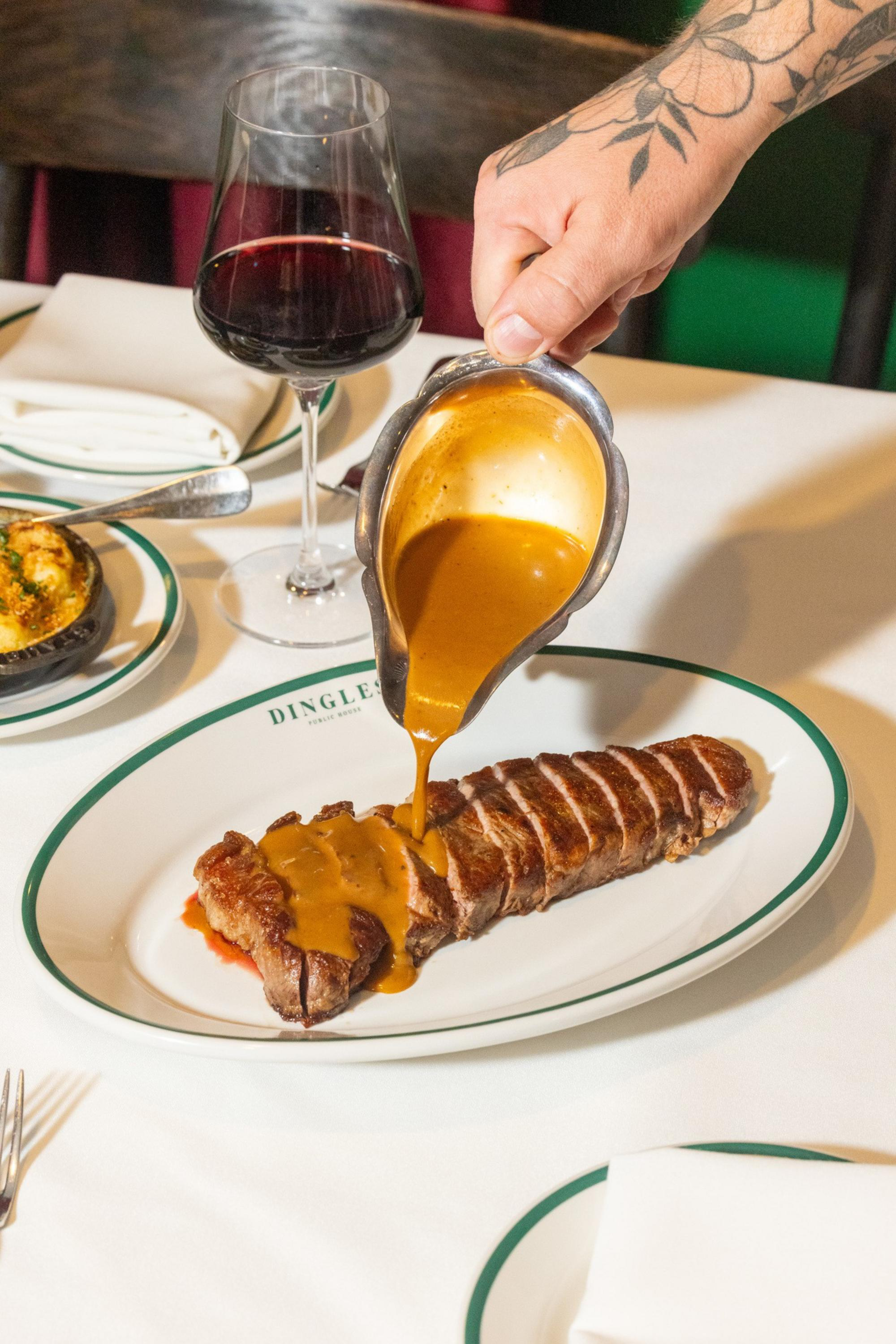 A hand with a tattoo pours brown sauce over a sliced steak on a white plate, with a glass of red wine and a side dish in the background.
