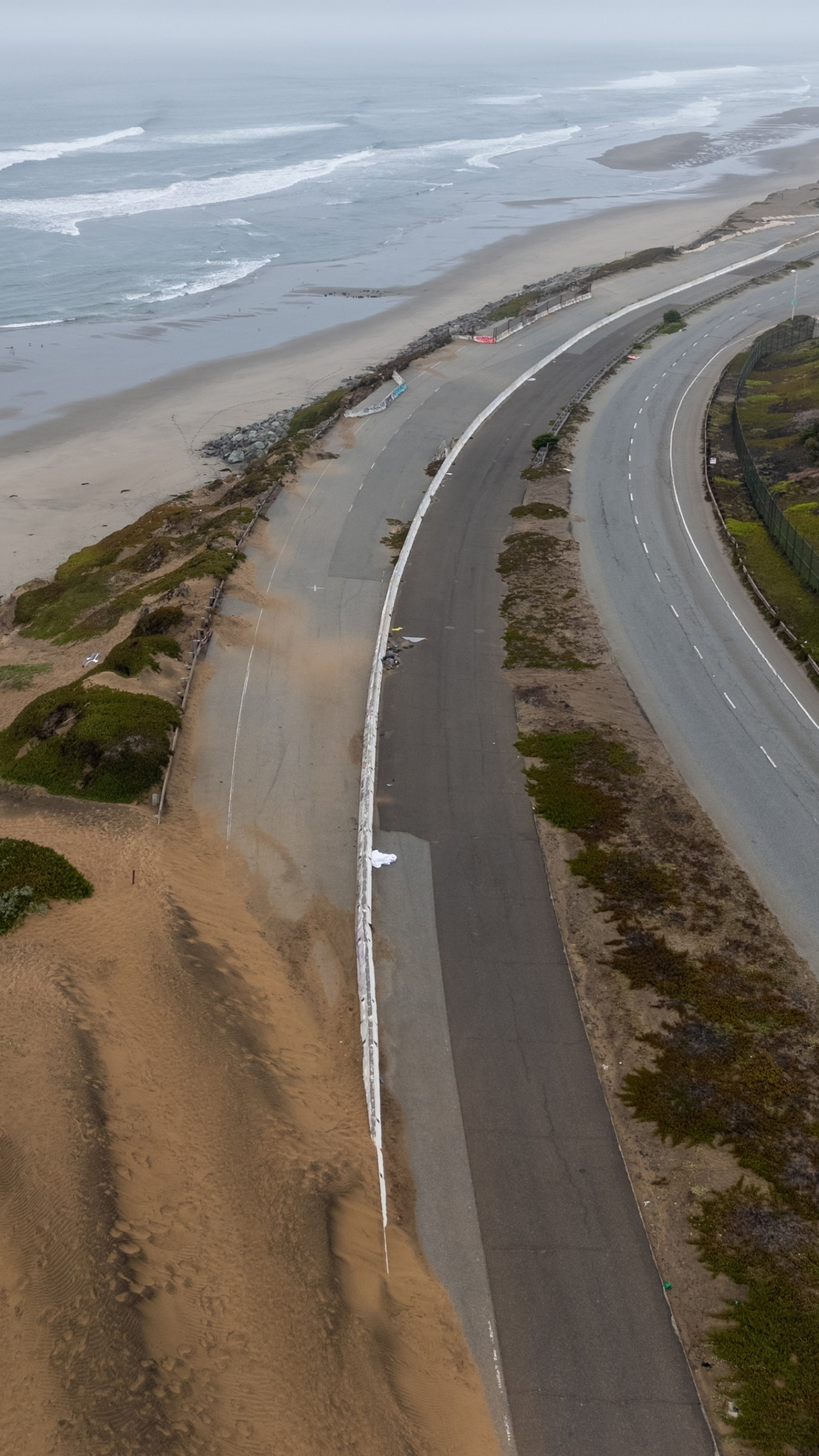 A coastal road is partially covered by sand dunes, with the ocean on one side and greenery on the other, under a cloudy sky.