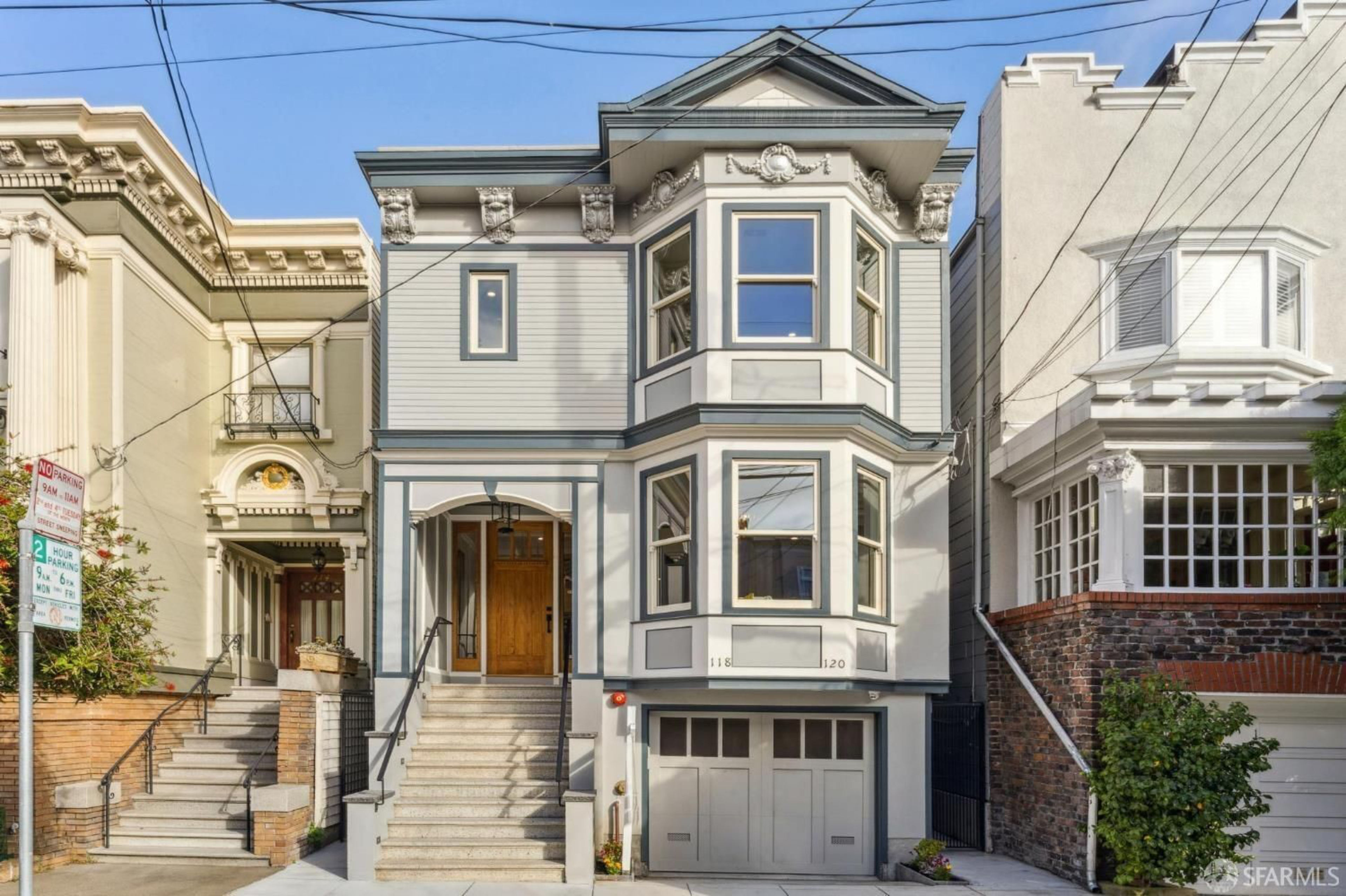 A three-story gray and white Victorian house with bay windows, a wooden front door, stairs leading to the entrance, and a garage door below.