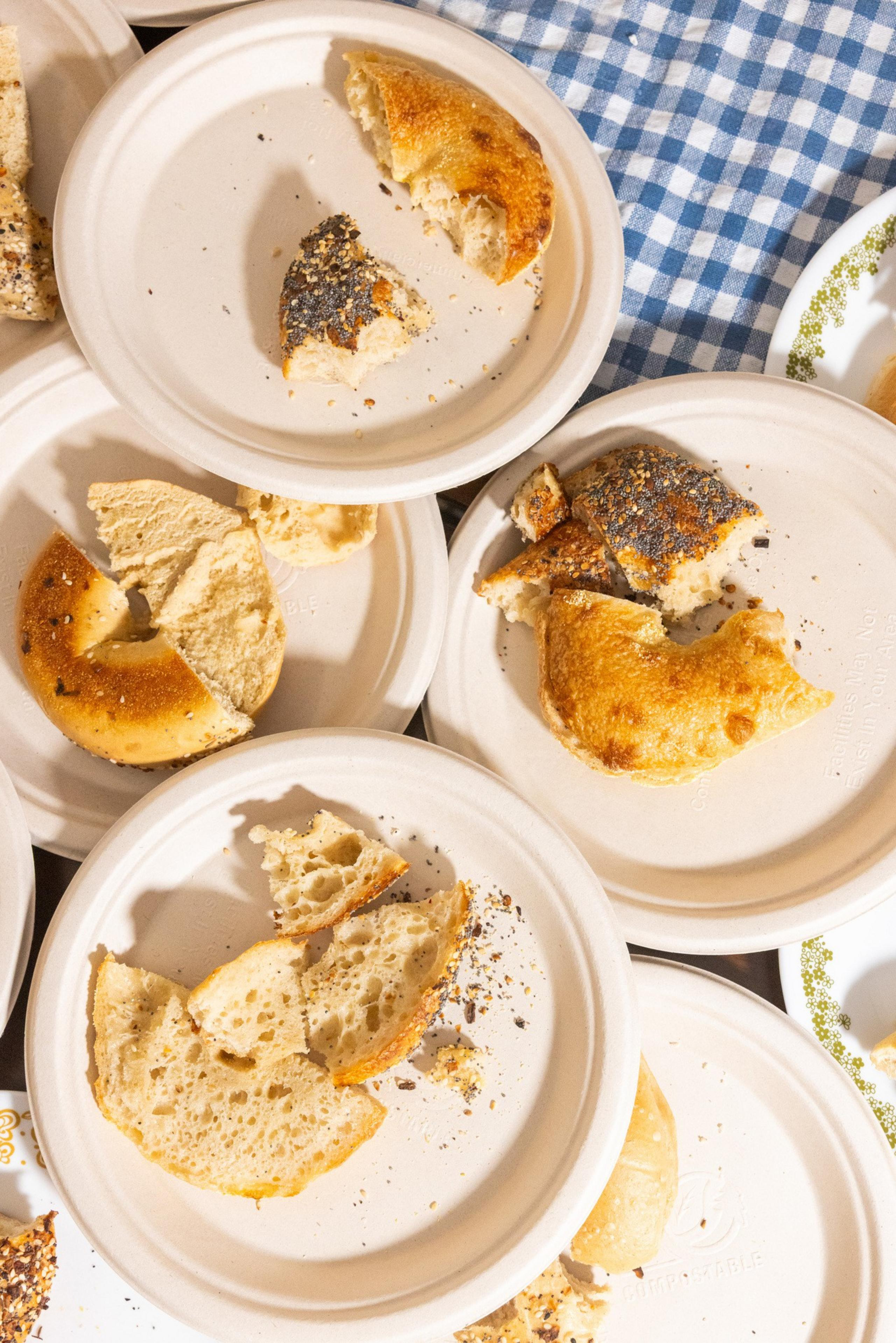 Several paper plates with partially eaten bagels sit atop a blue-checked cloth. Poppy seeds and crumbs are scattered around.