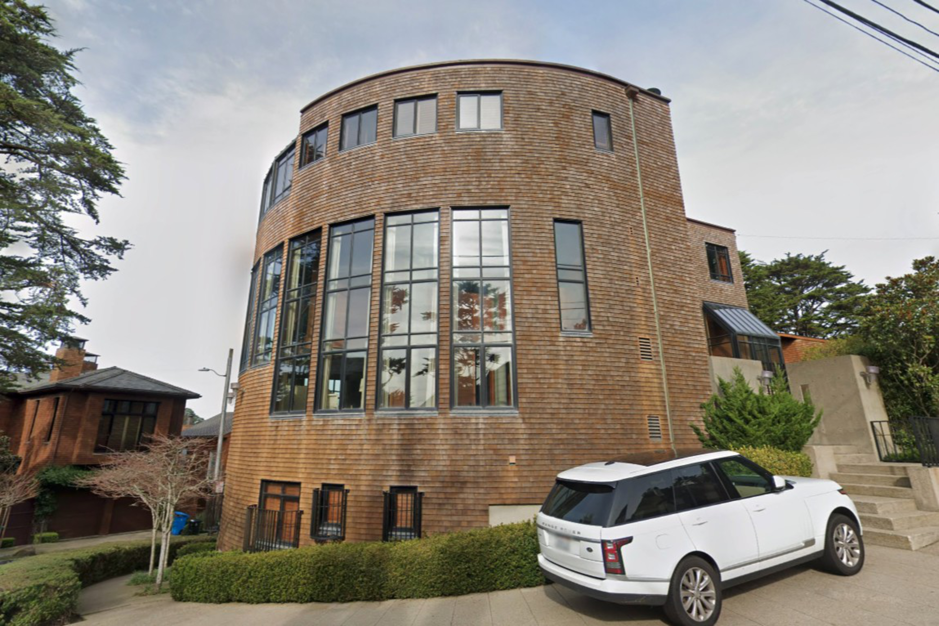 A round, multi-story house with wooden siding features large vertical windows, surrounded by greenery, with a white SUV parked on a sloped driveway.