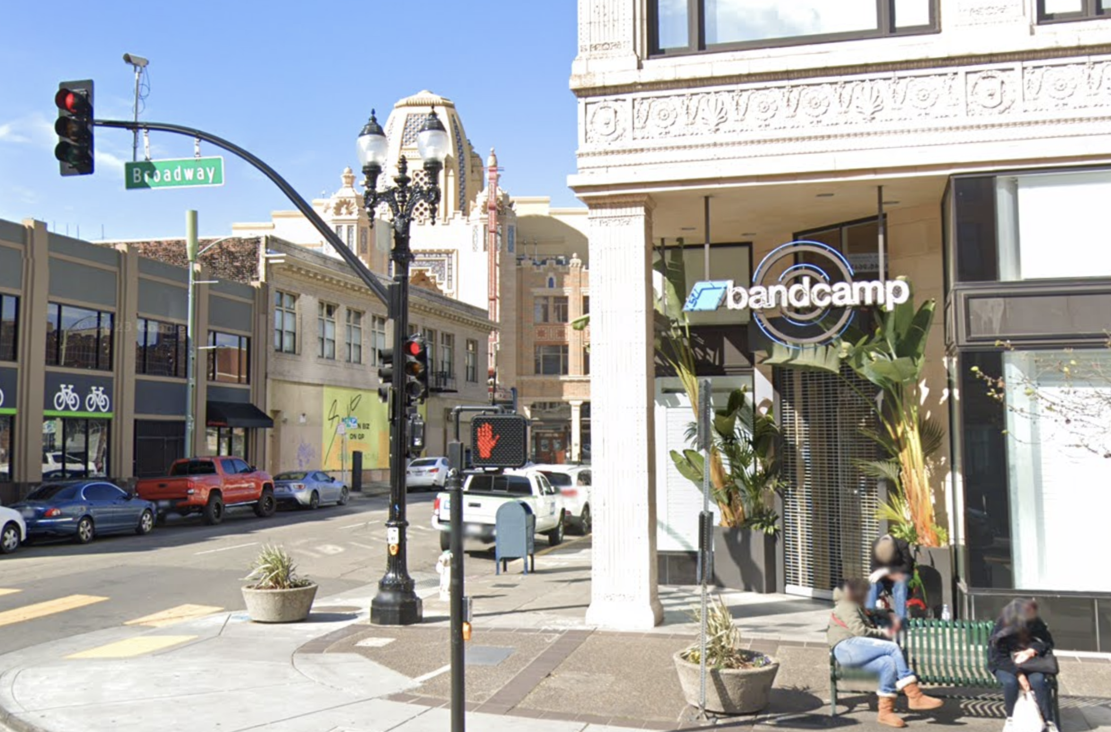 A sunny urban street corner with a Bandcamp office, people sitting on a bench, cars parked, and a Broadway street sign visible.