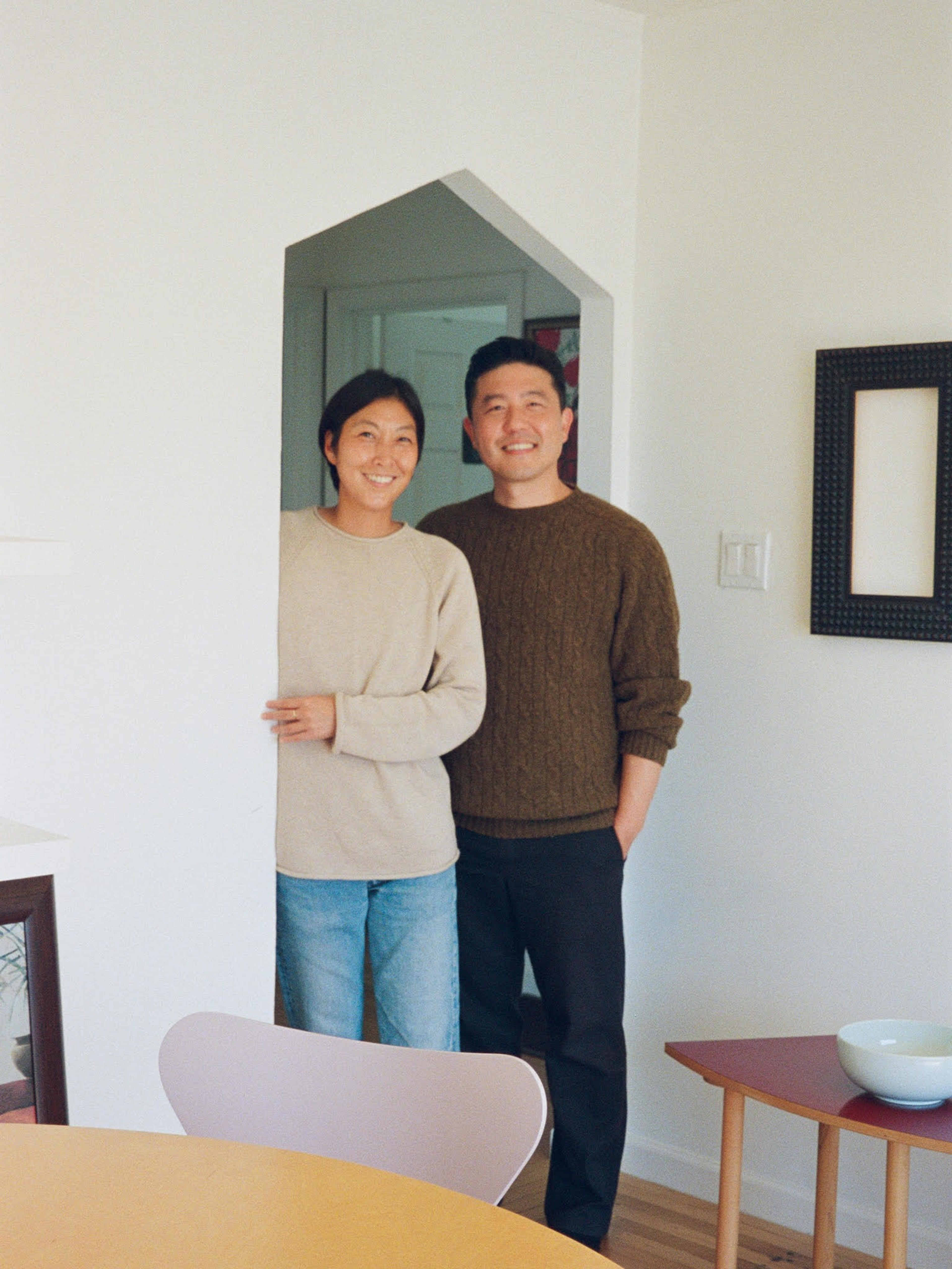A smiling couple stands in a doorway; the woman wears a beige sweater and jeans, and the man wears a brown sweater and black pants.