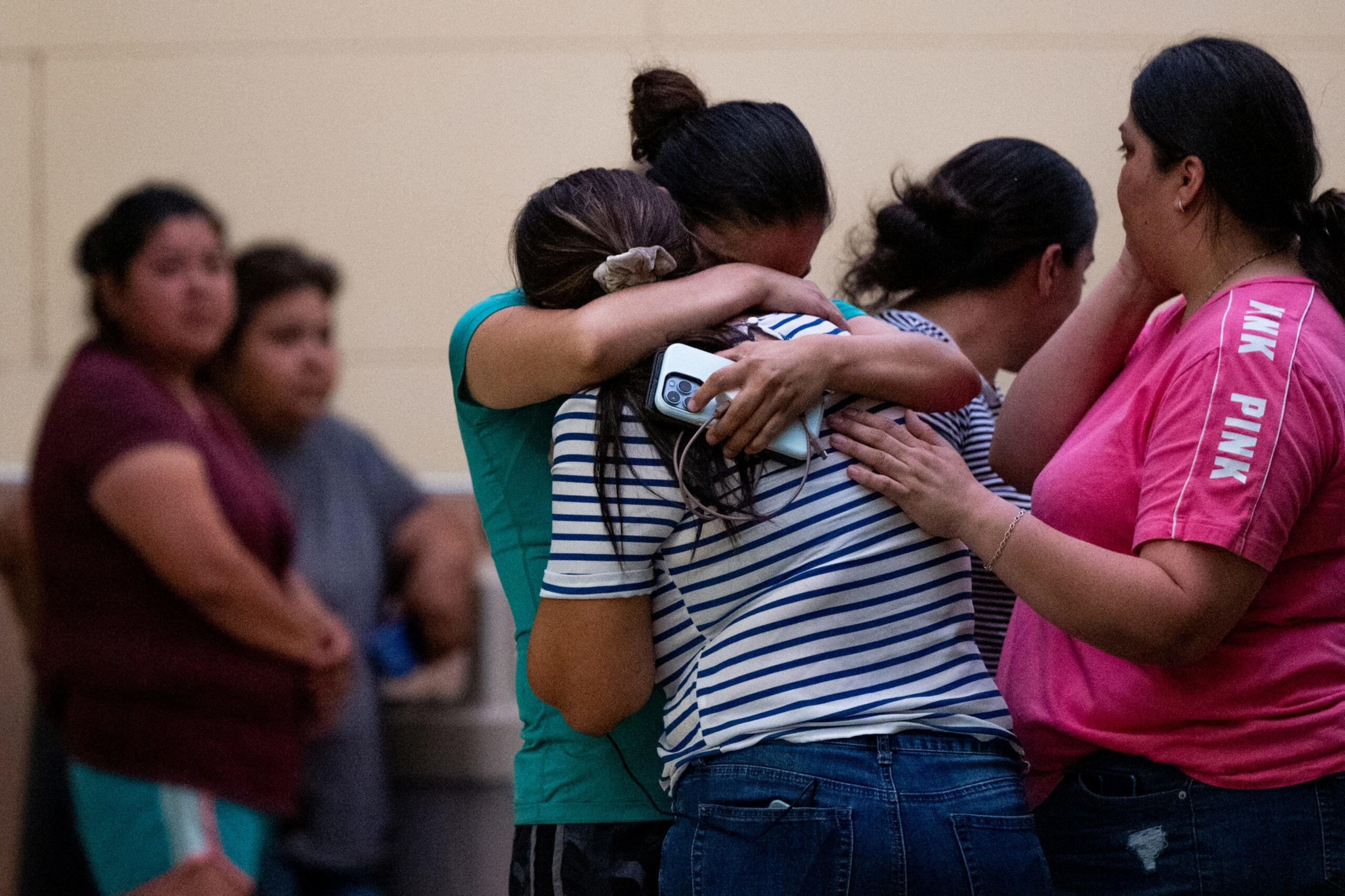 Four women are consoling each other, with two hugging tightly and another wiping tears, while two more stand somberly in the background.