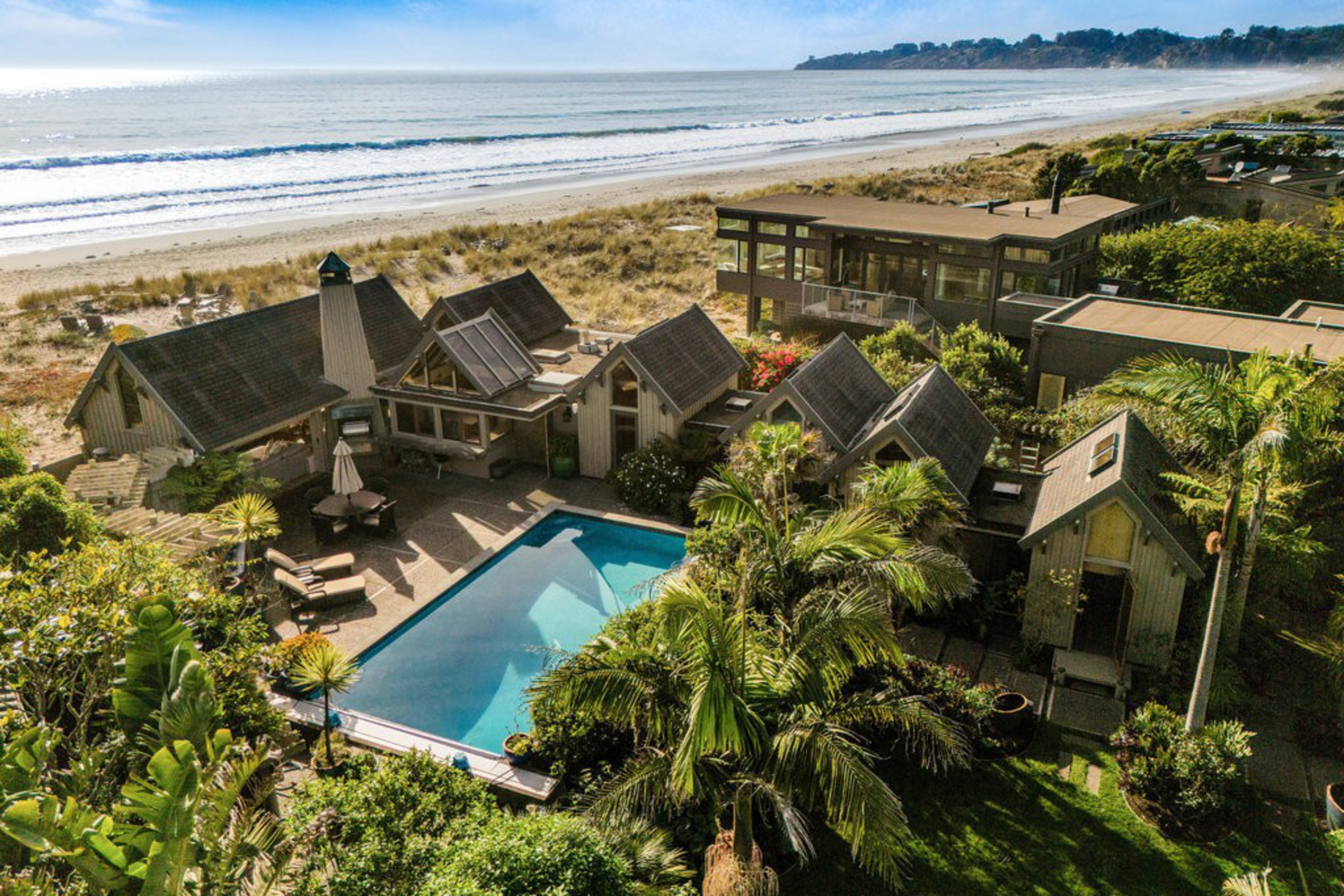 A beachside property with multiple small buildings surrounds a rectangular swimming pool, with palm trees and a sandy shore leading to the ocean.