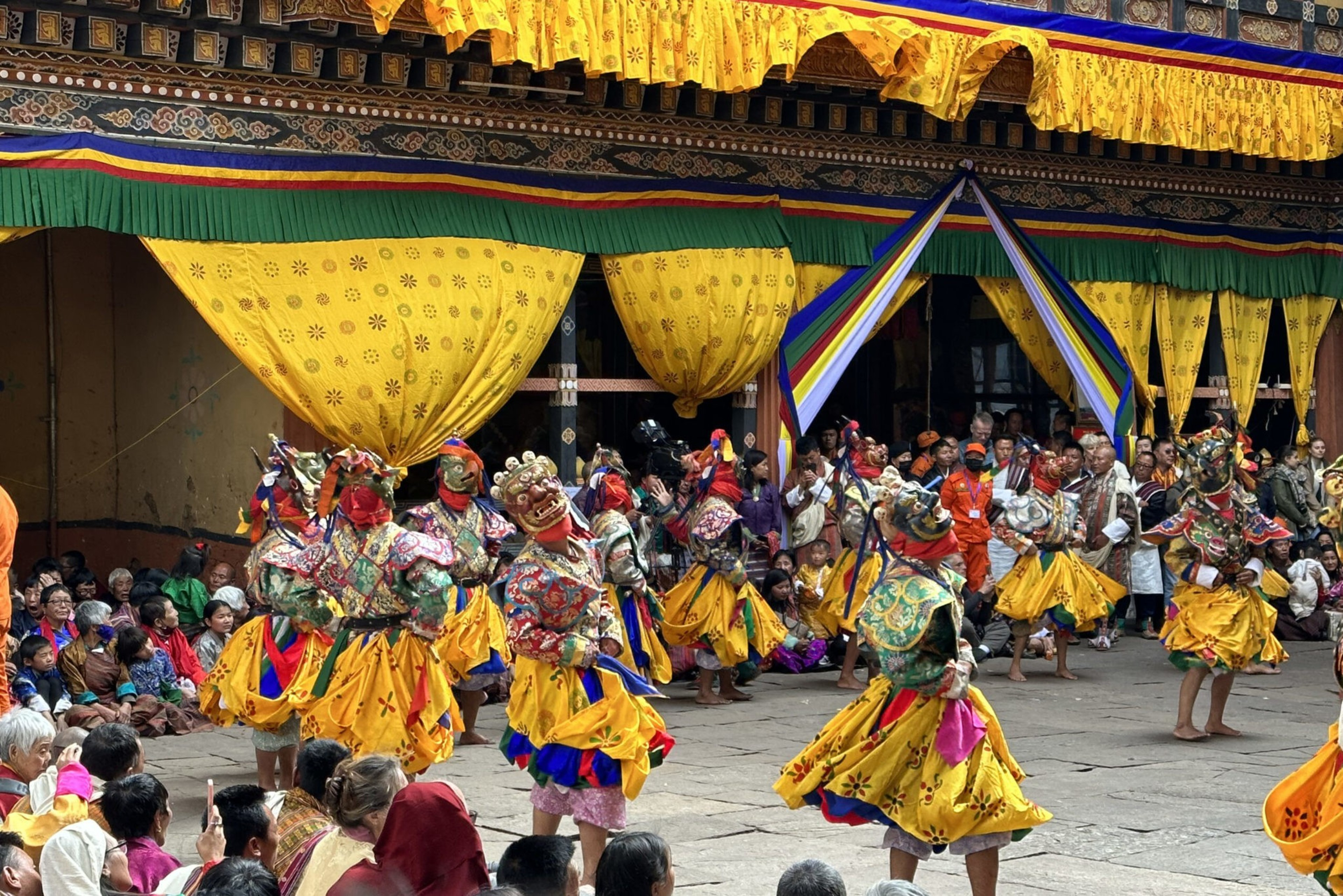 Dancers wearing colorful traditional costumes and masks perform in front of an audience under yellow and multi-colored decorated canopy.
