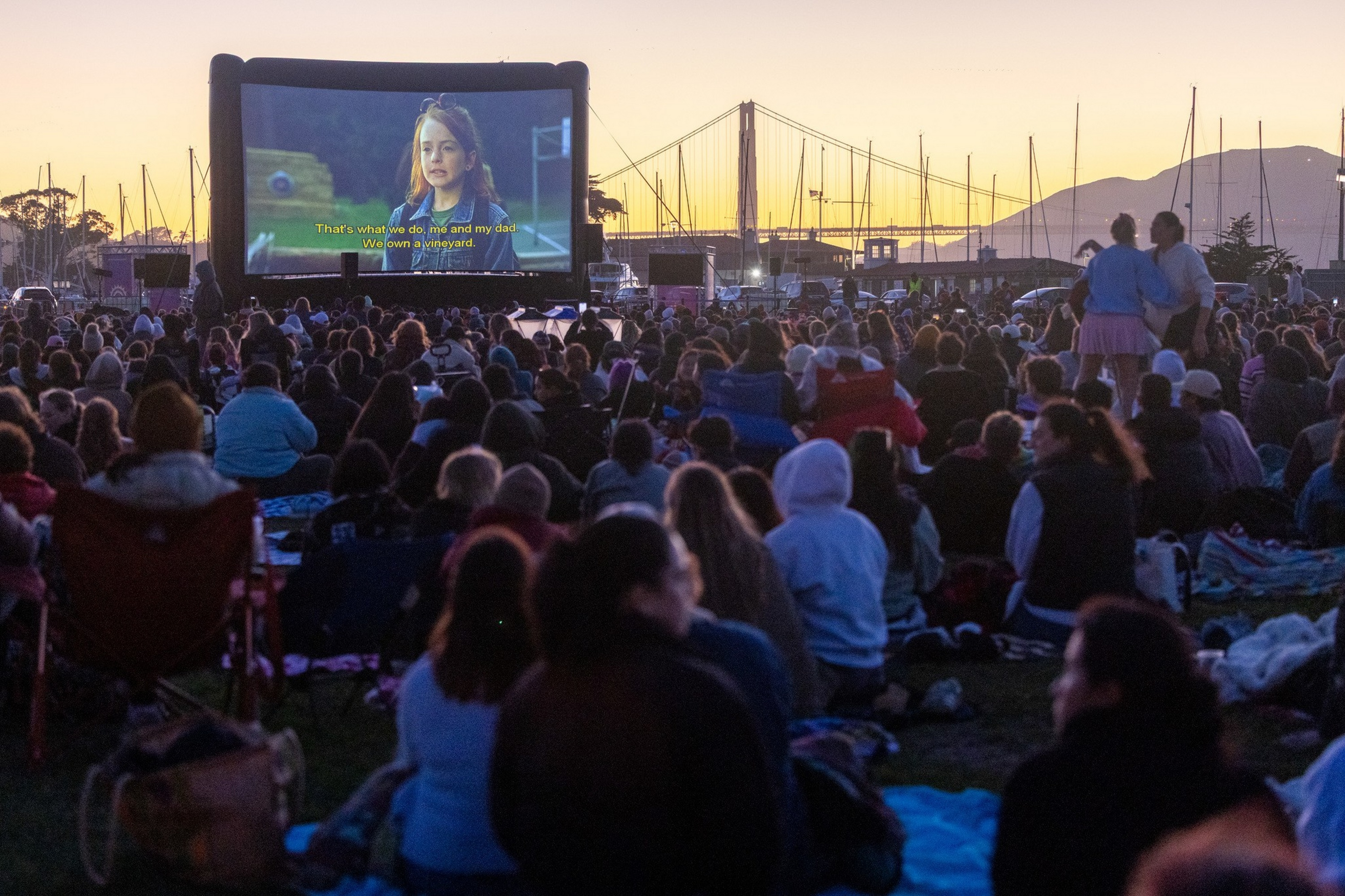 A large crowd watches a movie on an outdoor screen at dusk near a marina with boats and a bridge silhouetted against the sunset.
