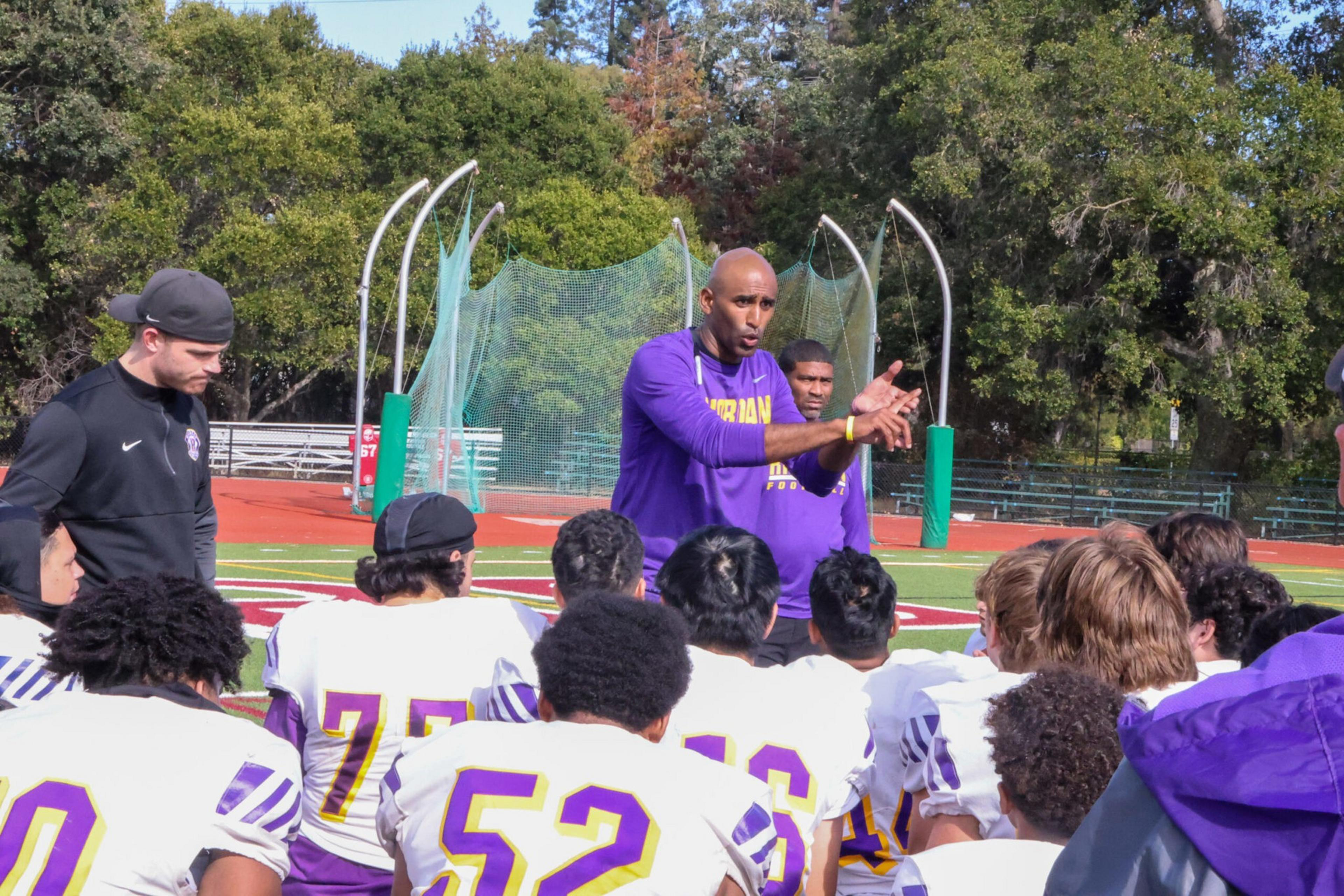 A coach in a purple shirt talks passionately to a group of football players wearing white and purple uniforms on a sunny field.