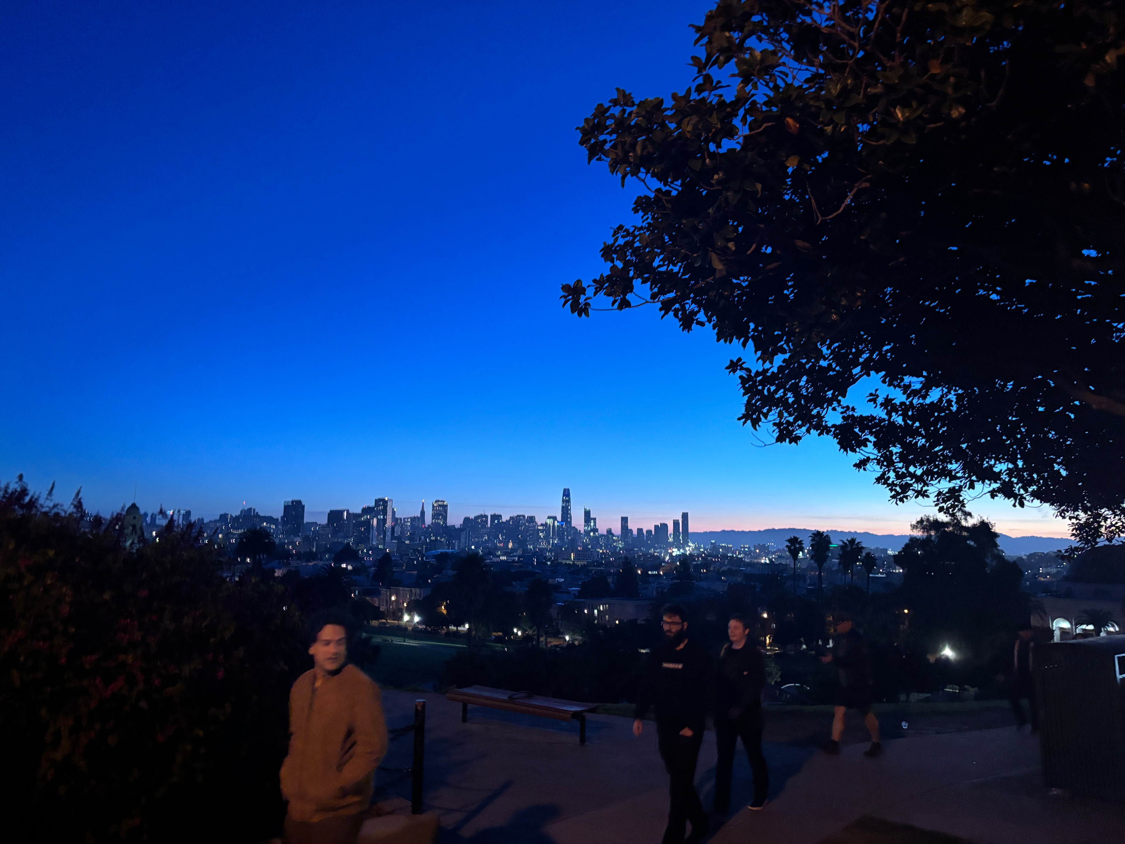 A city skyline glows under a deep blue twilight sky with several people walking along a park path framed by dark trees.