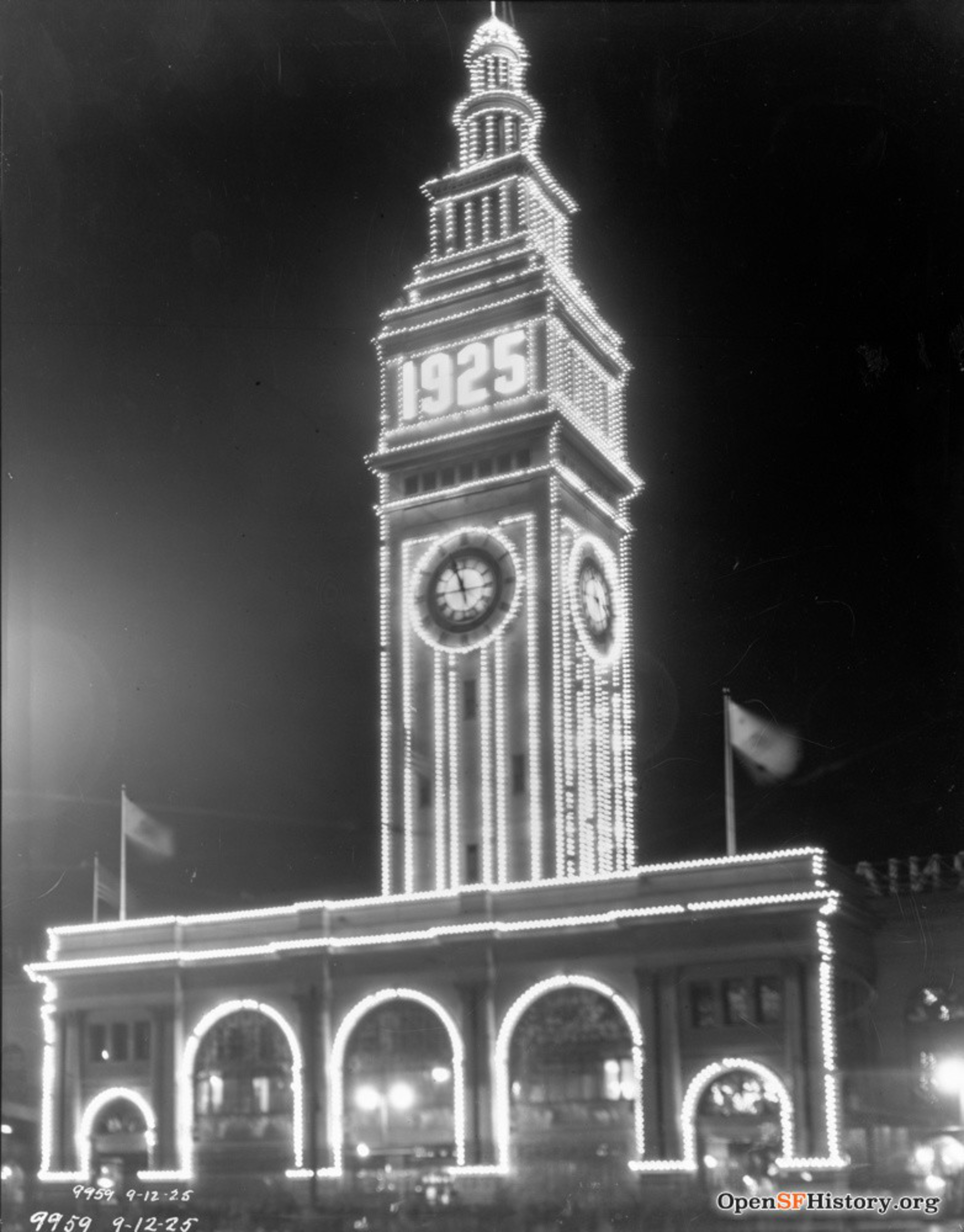 A tall clock tower outlined in bright lights shows "1925" near the top, with the time on the clock faces reading about 8:30. Flags fly on the building below.