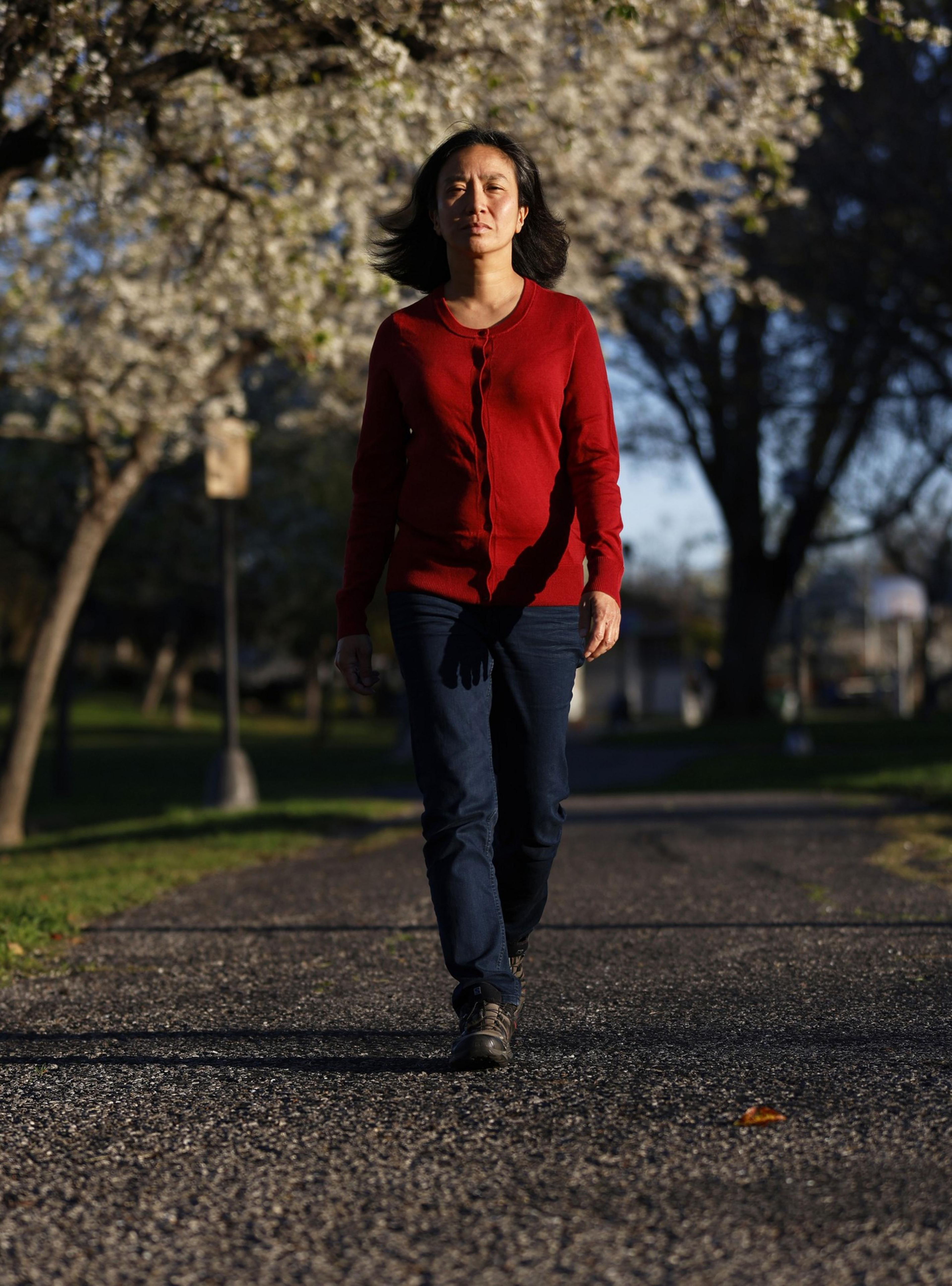 A person walks down a sunlit path lined with blossoming trees. They wear a red sweater and jeans, with the light casting shadows around them.