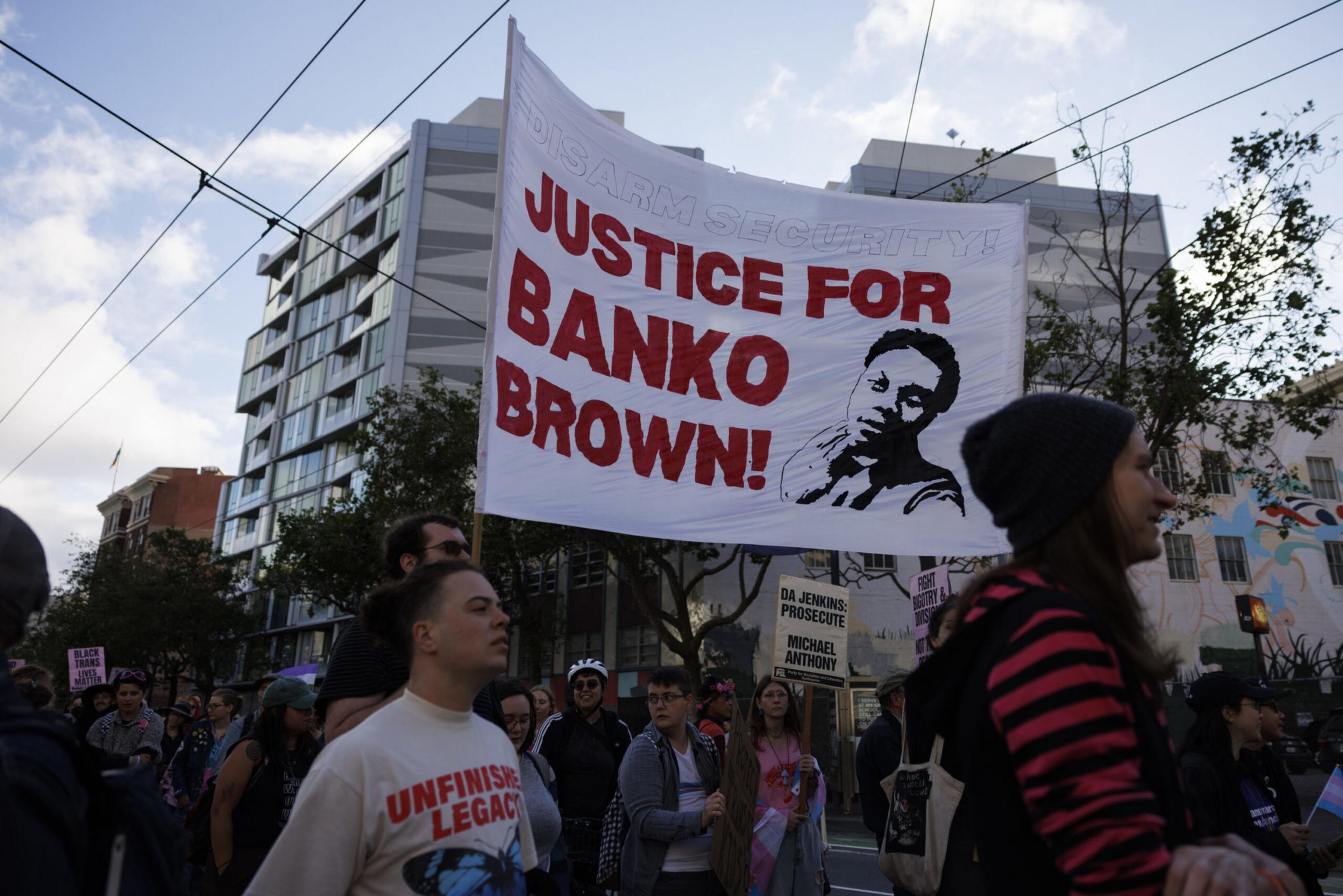 A group of protesters hold a large white banner reading "Justice for Banko Brown!" with an illustrated portrait in red and black.