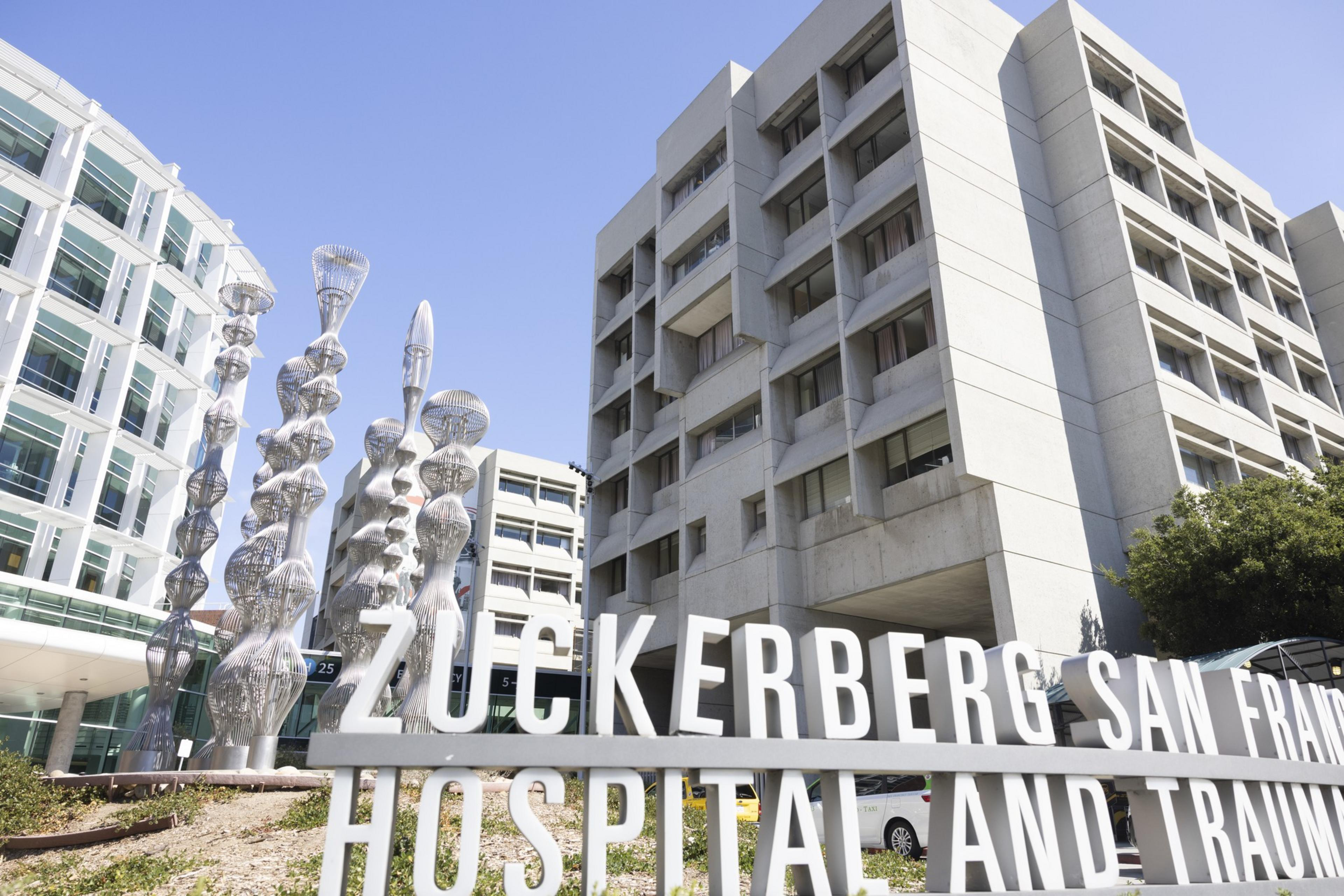 A hospital building with modern sculptures and a sign that reads &quot;Zuckerberg San Francisco Hospital and Trauma Center&quot; is shown under a clear blue sky.