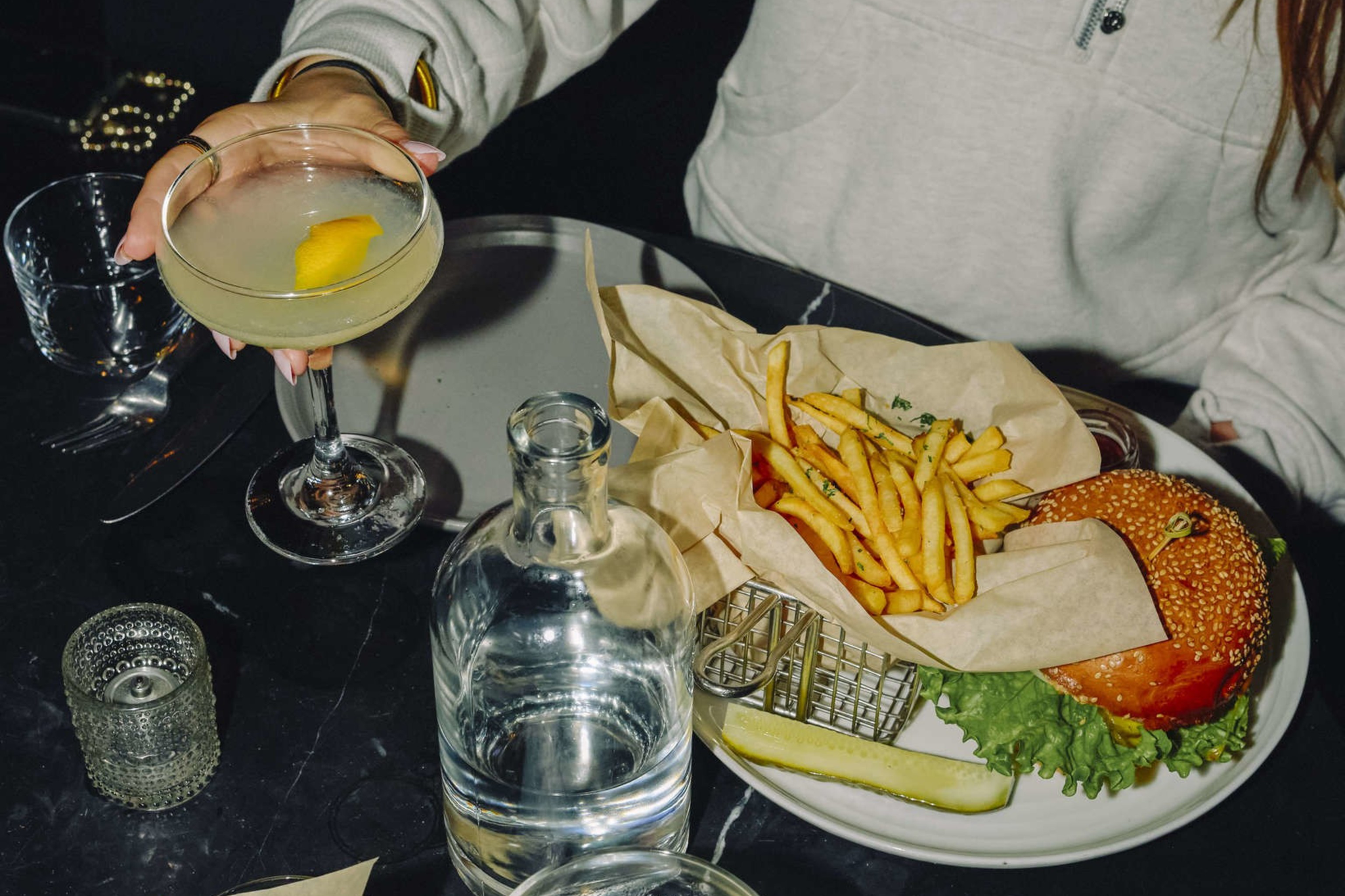 A person in a light zip-up sweater holds a cocktail glass with a lemon twist, with a plate of a burger, fries, and a pickle on the table.