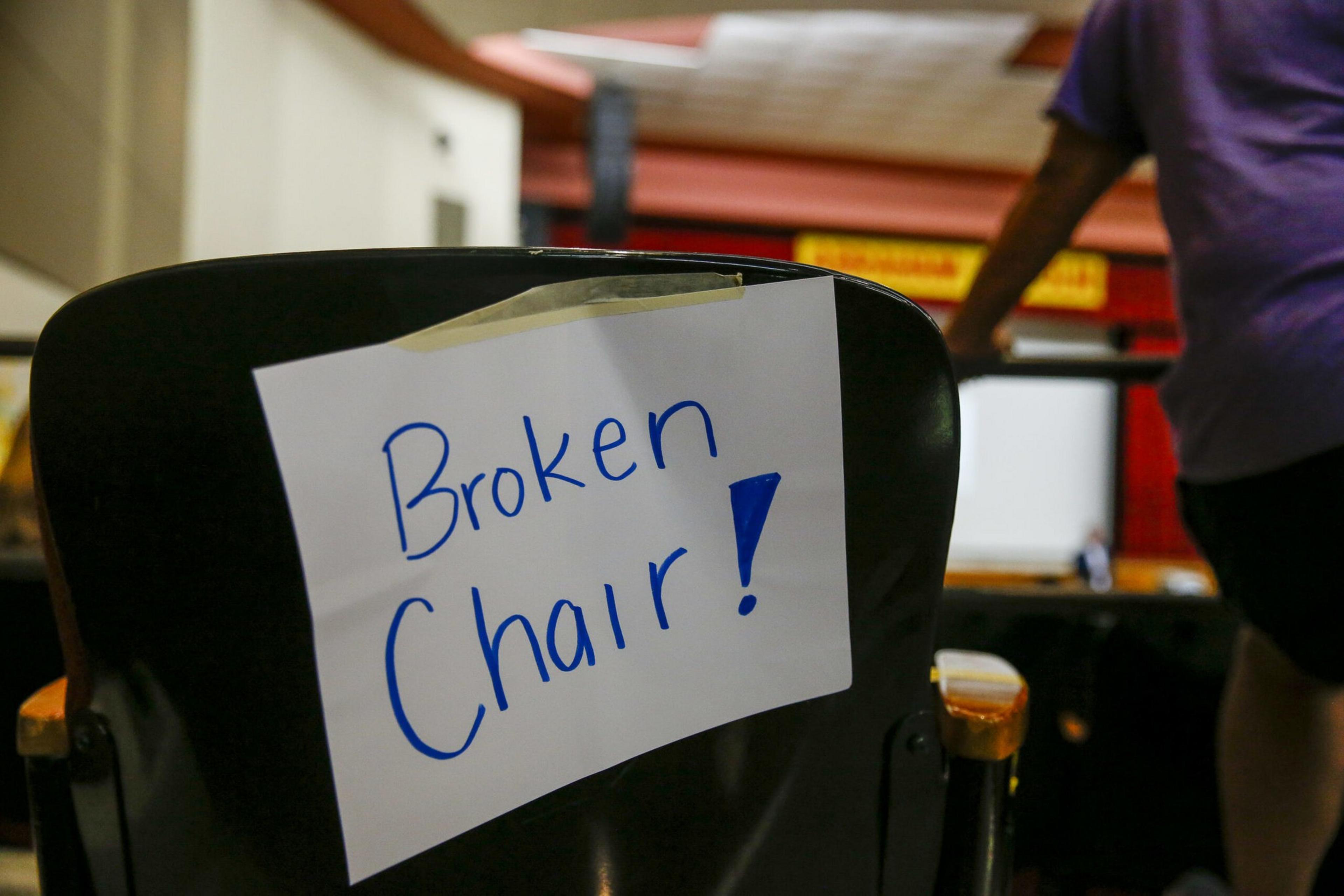 A black chair with wooden armrests has a taped white paper sign reading "Broken Chair!" in blue marker. A person in a purple shirt stands nearby.