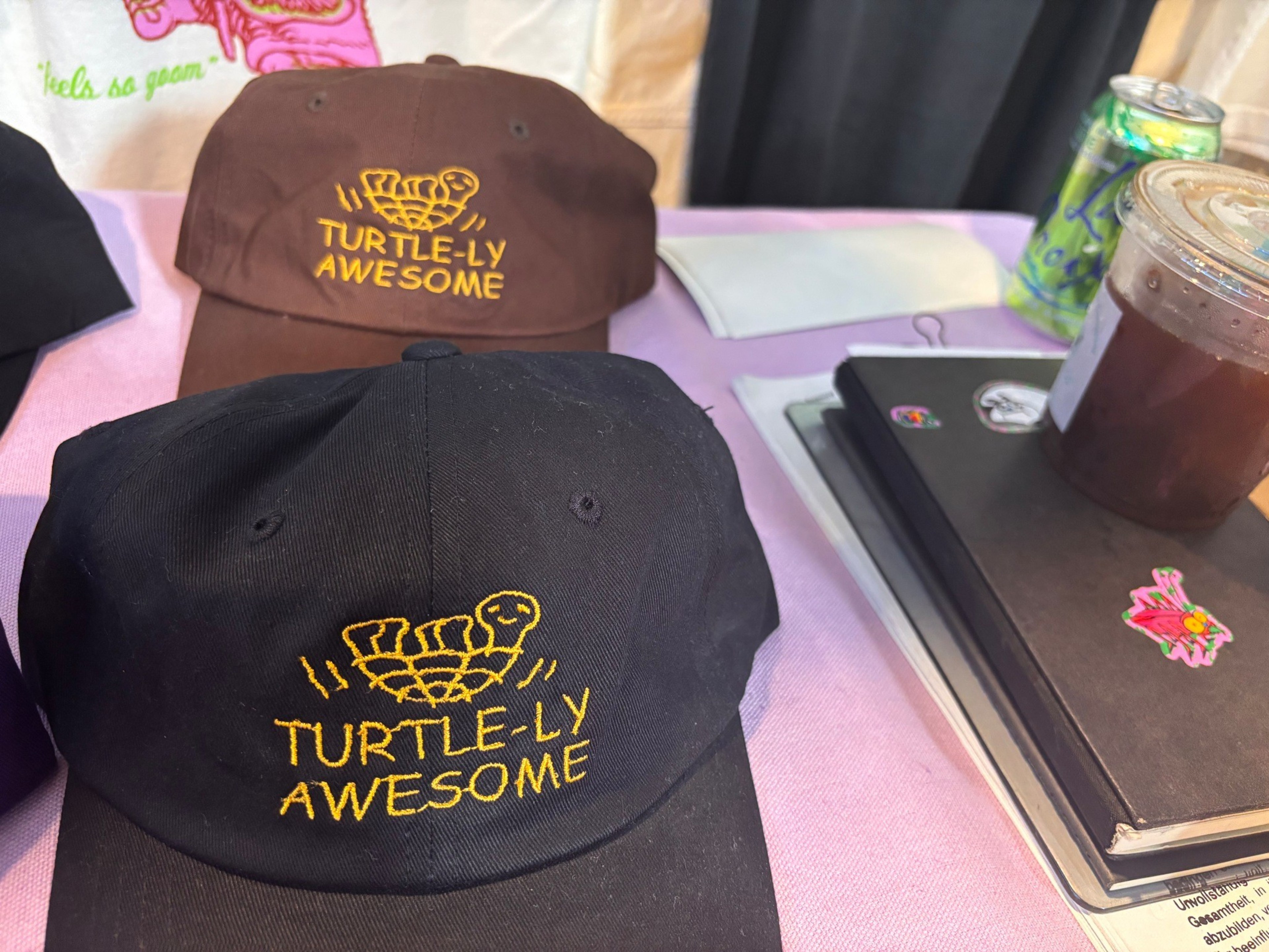 Two baseball caps, one black and one brown, each embroidered with a turtle and the phrase “TURTLE-LY AWESOME,” sit on a table next to notebooks and drinks.