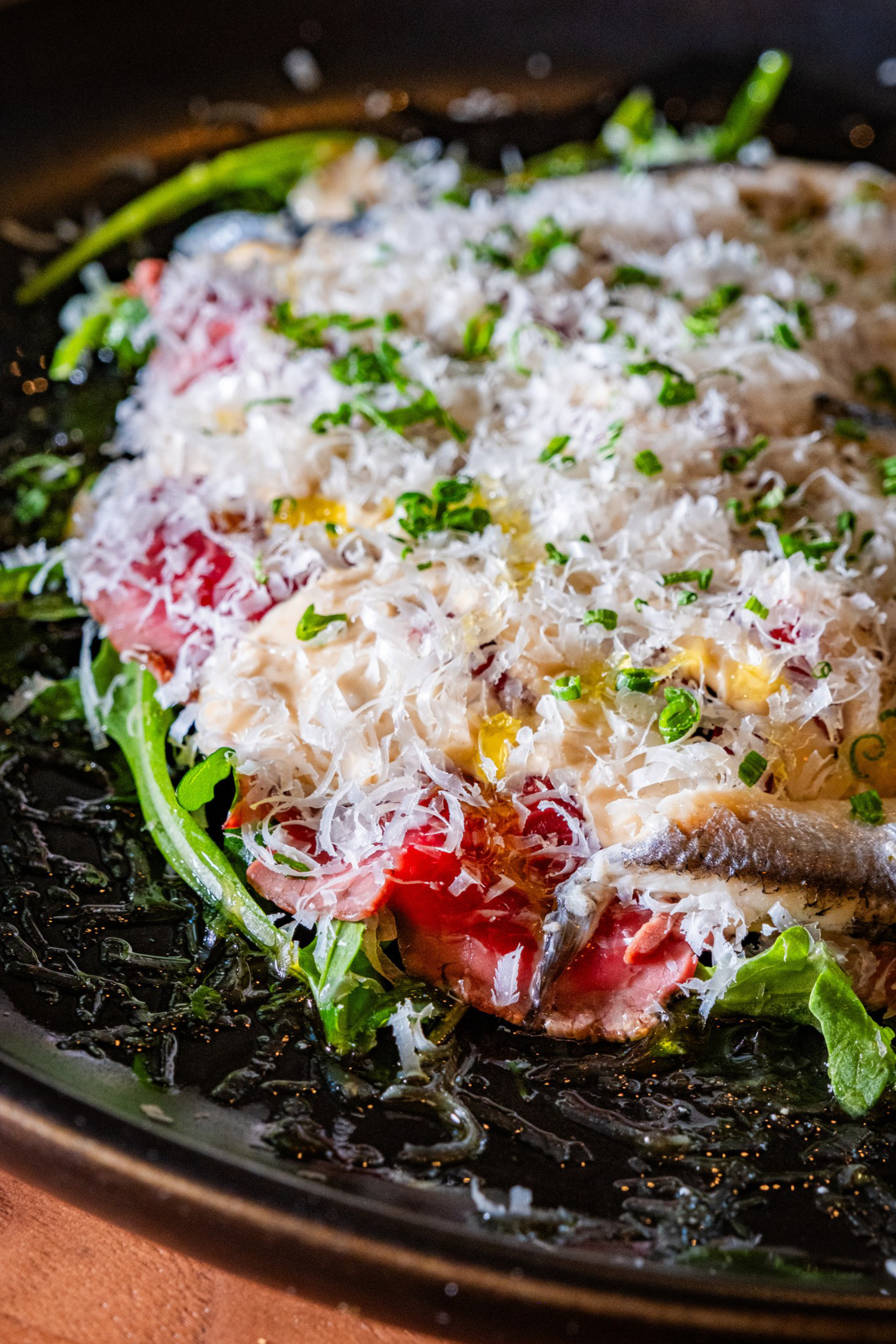 The image shows a dish with thinly sliced meat, topped with grated cheese, fresh greens, and garnished with herbs, all drizzled with olive oil.