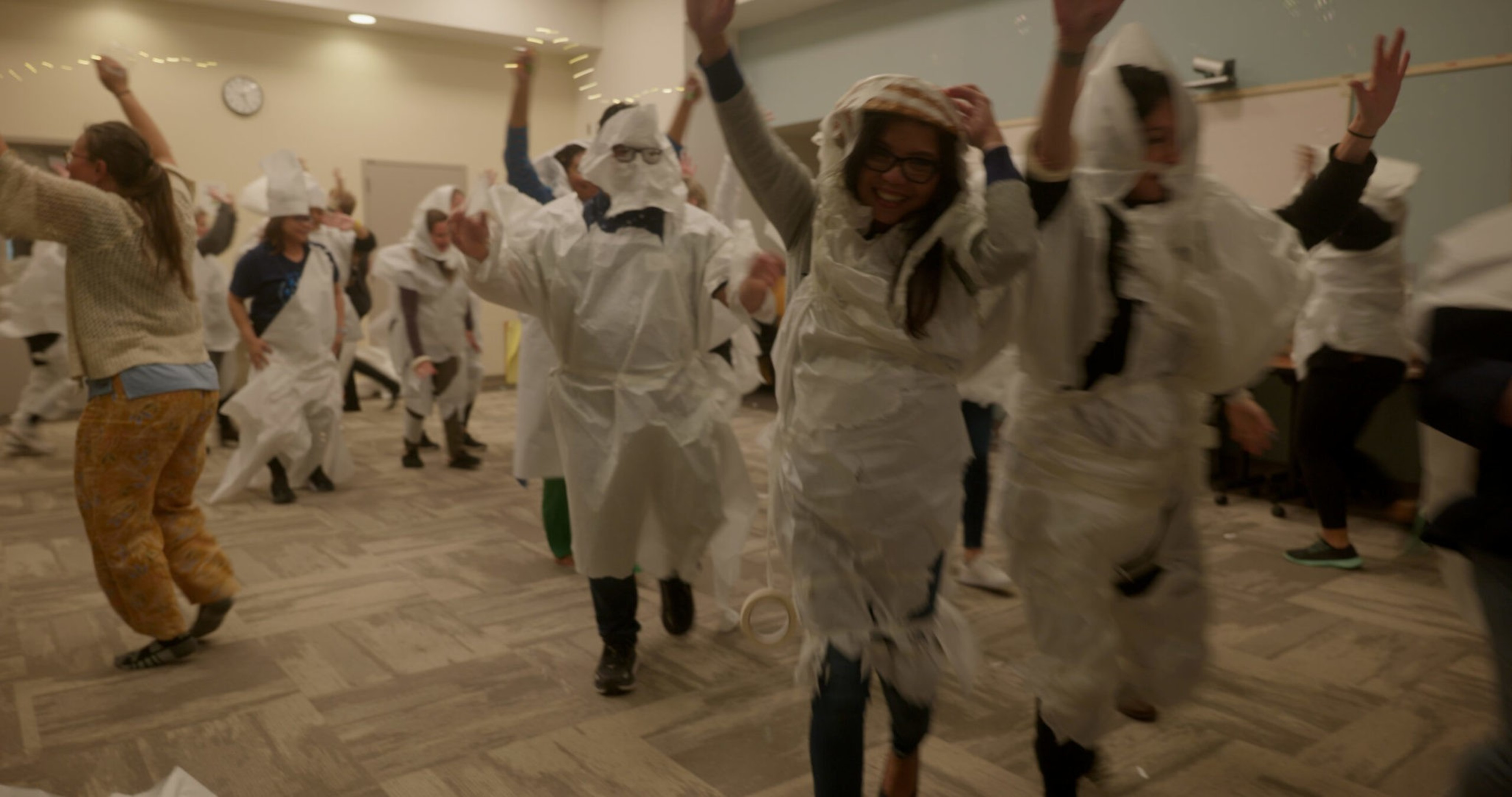 People wearing costumes made of white paper or plastic, dancing and having fun in a brightly lit indoor room.