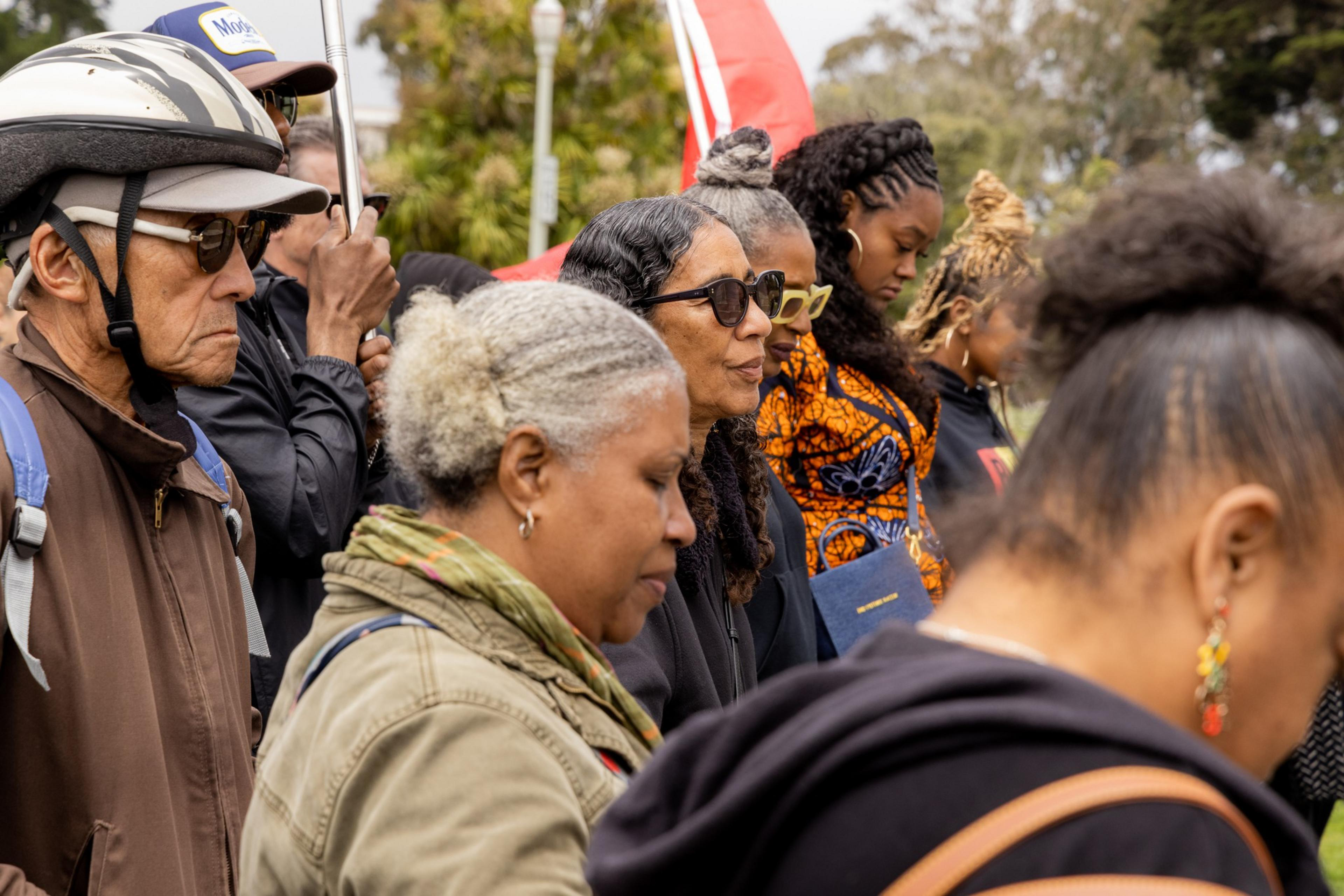 A group of people, including men and women with varied hairstyles and accessories, are gathered outdoors, seemingly in a solemn or attentive moment, with a red flag in the background.