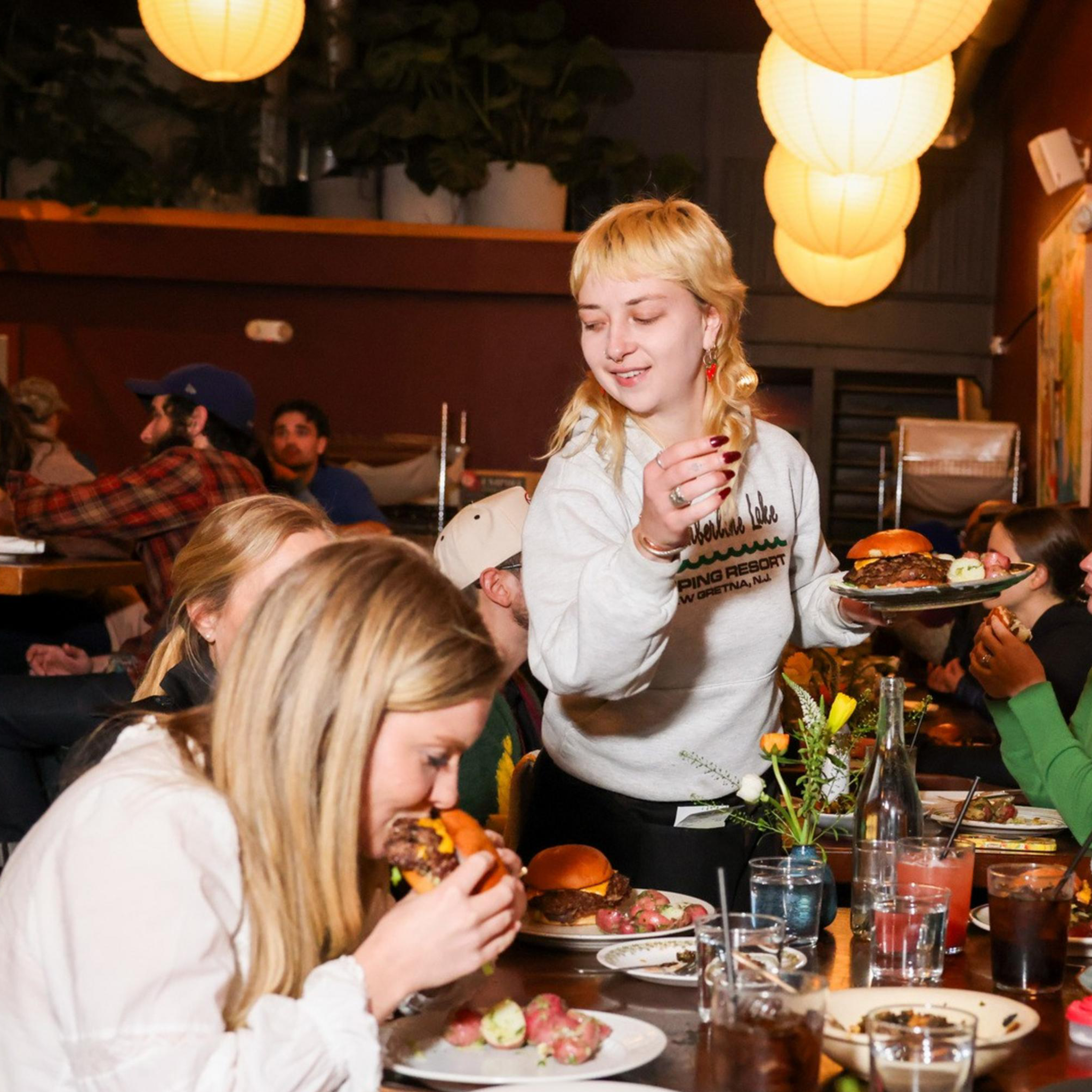 A cozy restaurant scene with people enjoying burgers and drinks. A person in white is serving food, while others take photos and chat around a wooden table.