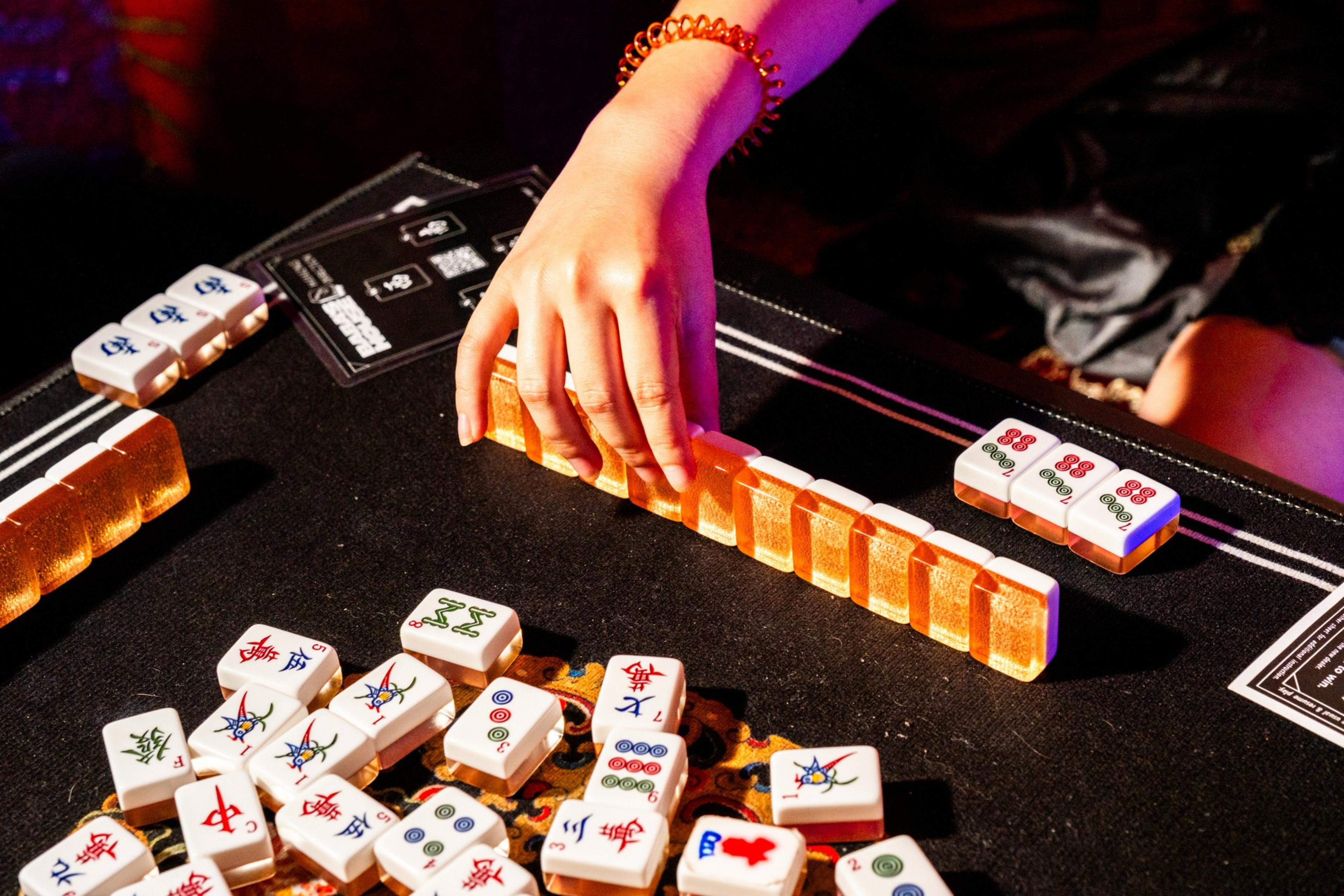 A person with a beaded bracelet adjusts a row of vibrant mahjong tiles on a dark table, surrounded by scattered tiles, under warm lighting.