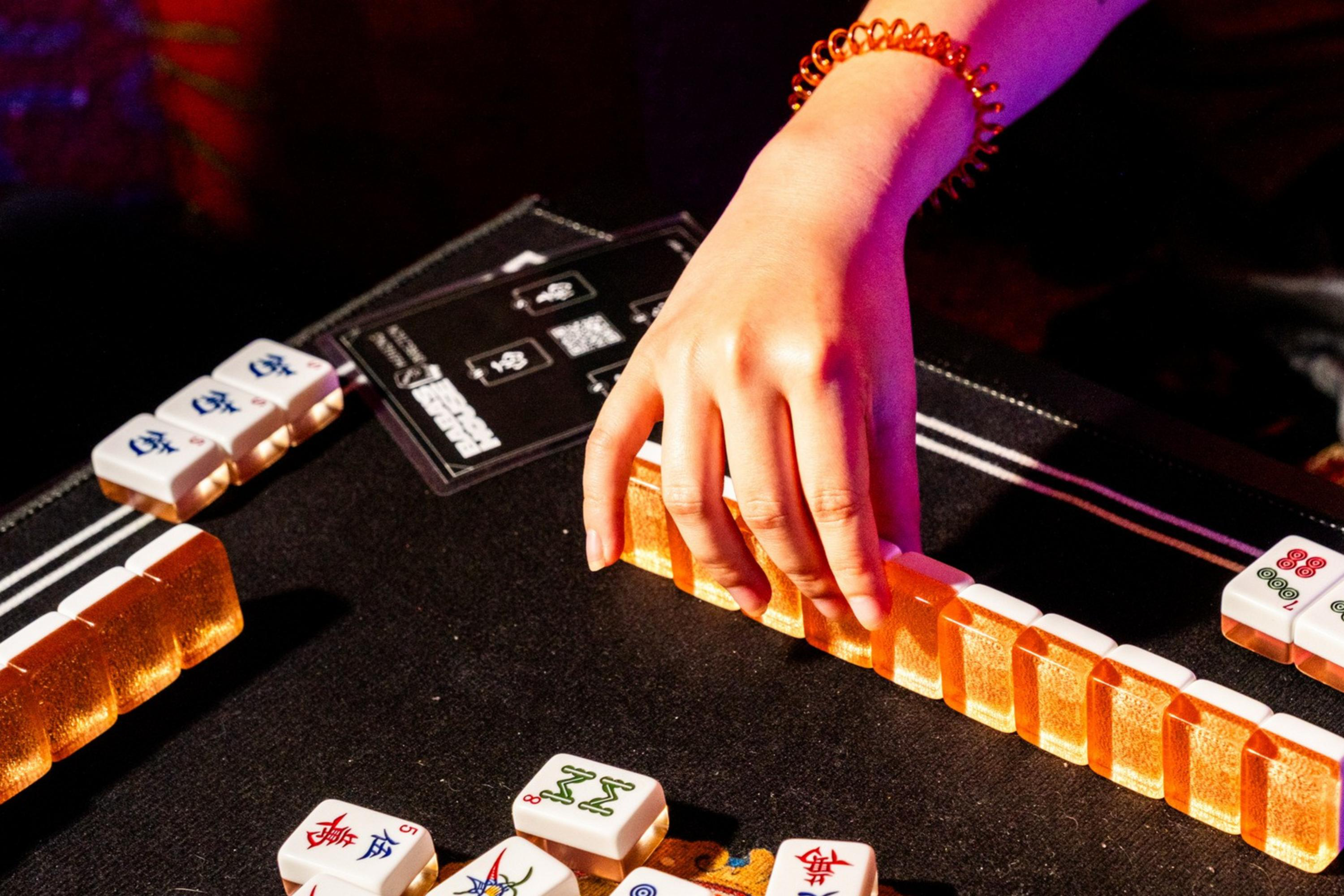 A person with a beaded bracelet adjusts a row of vibrant mahjong tiles on a dark table, surrounded by scattered tiles, under warm lighting.