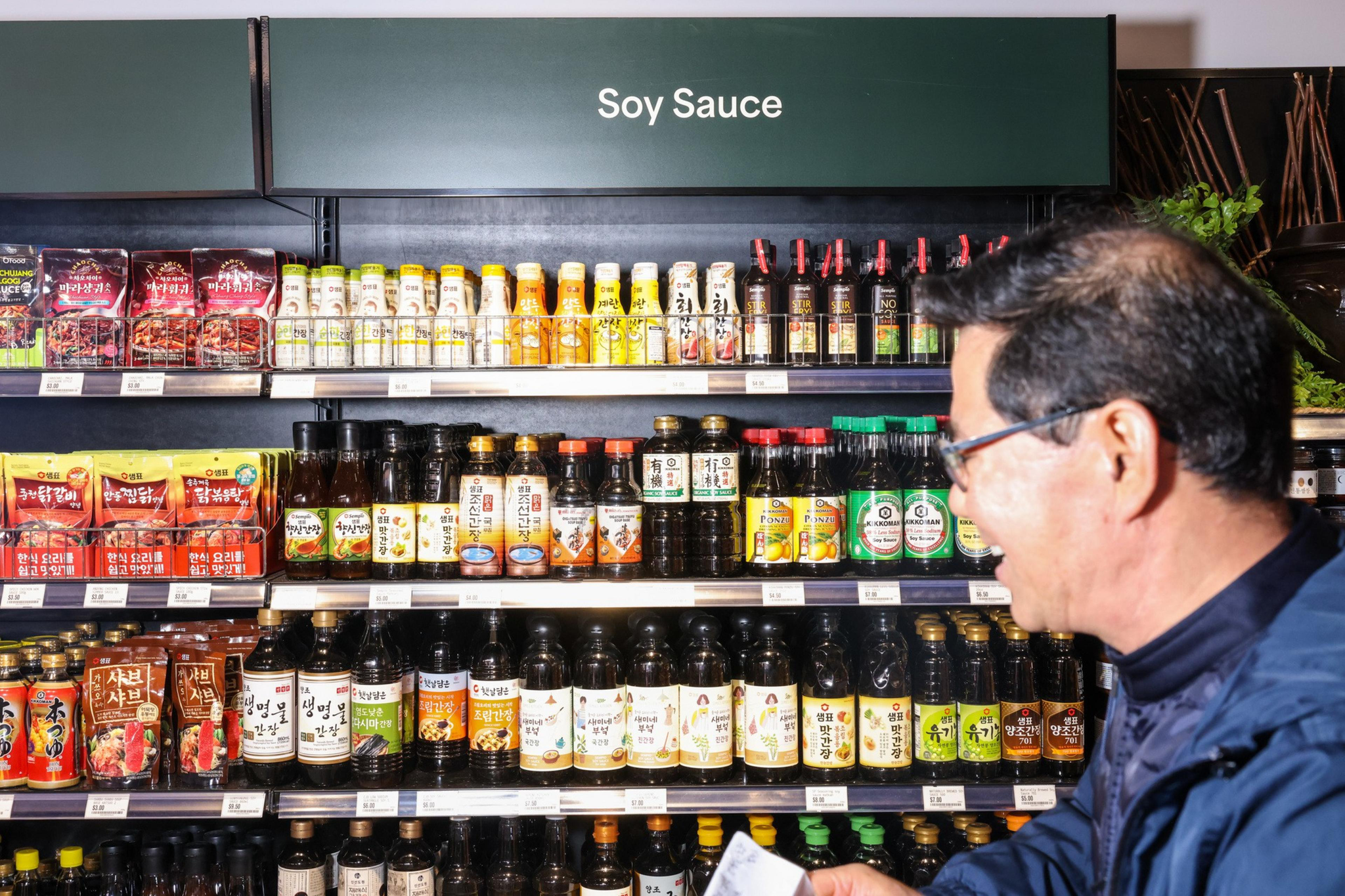 A man stands in front of shelves filled with various soy sauce bottles and condiments in a store, under a sign labeled "Soy Sauce".