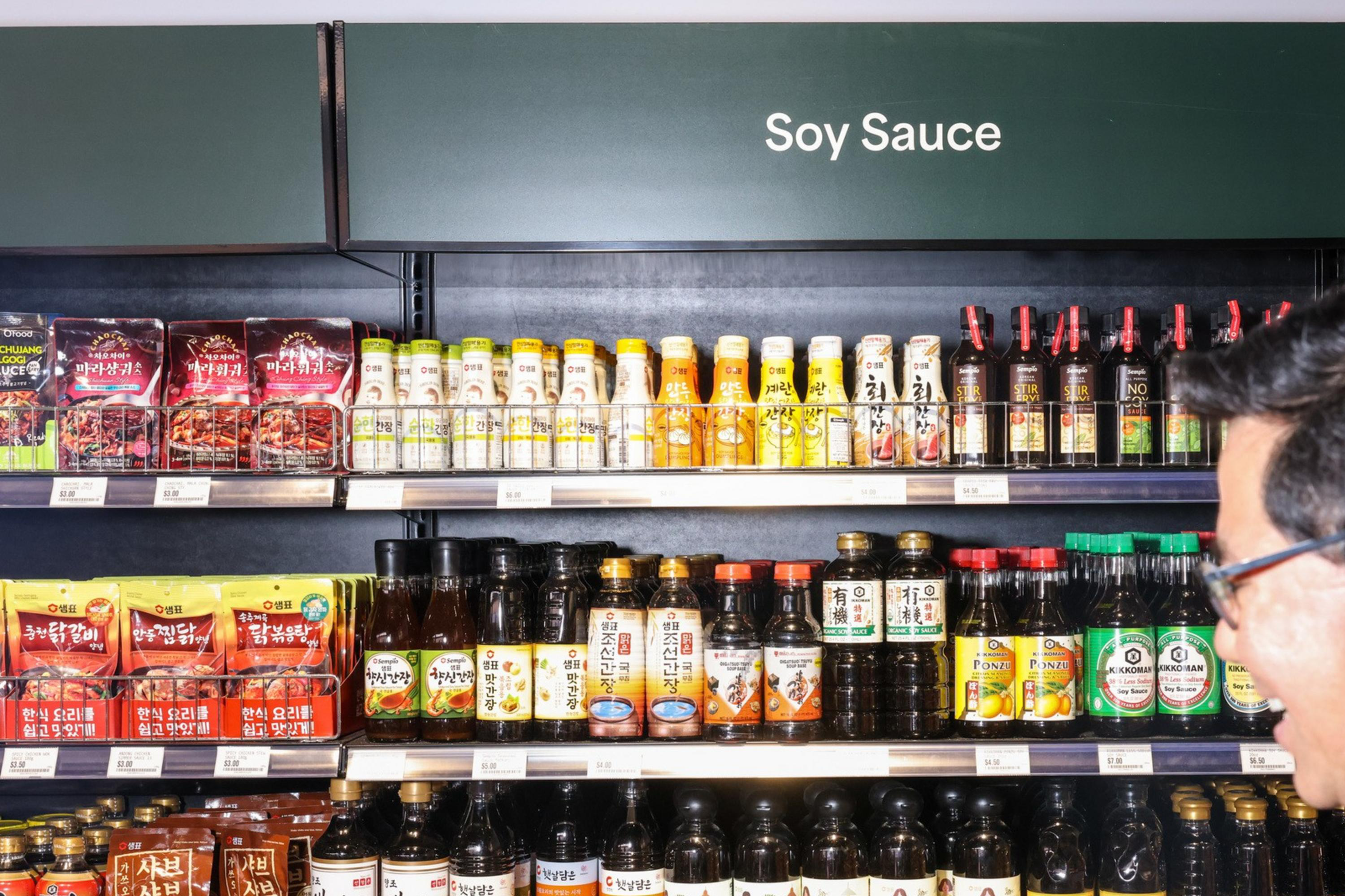A man stands in front of shelves filled with various soy sauce bottles and condiments in a store, under a sign labeled "Soy Sauce".