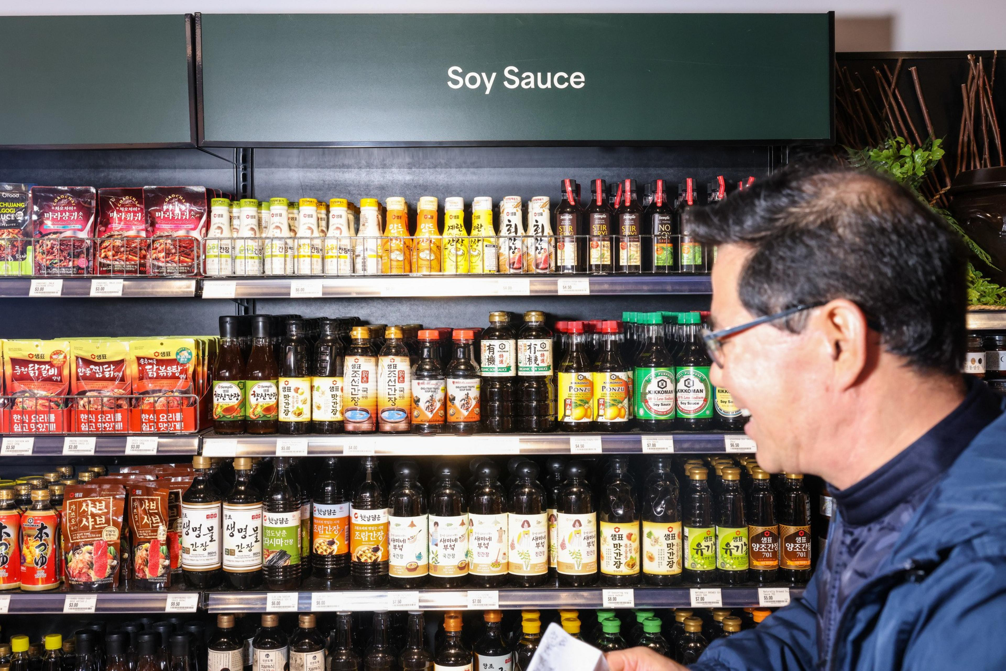 A man stands in front of shelves filled with various soy sauce bottles and condiments in a store, under a sign labeled "Soy Sauce".