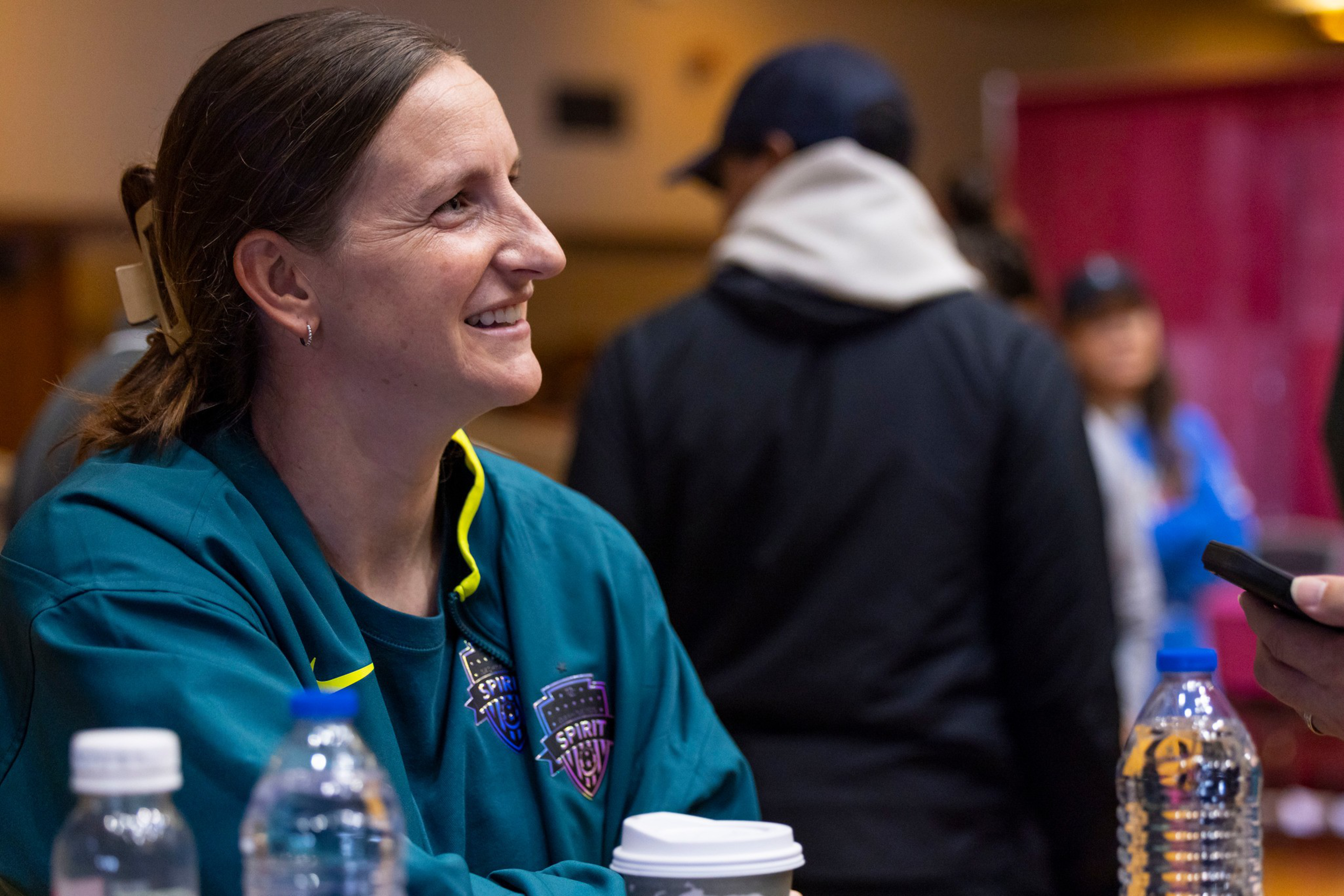 A woman wearing a teal jacket with a “Spirit” logo smiles while sitting at a table with water bottles and a coffee cup, people blurred in the background.