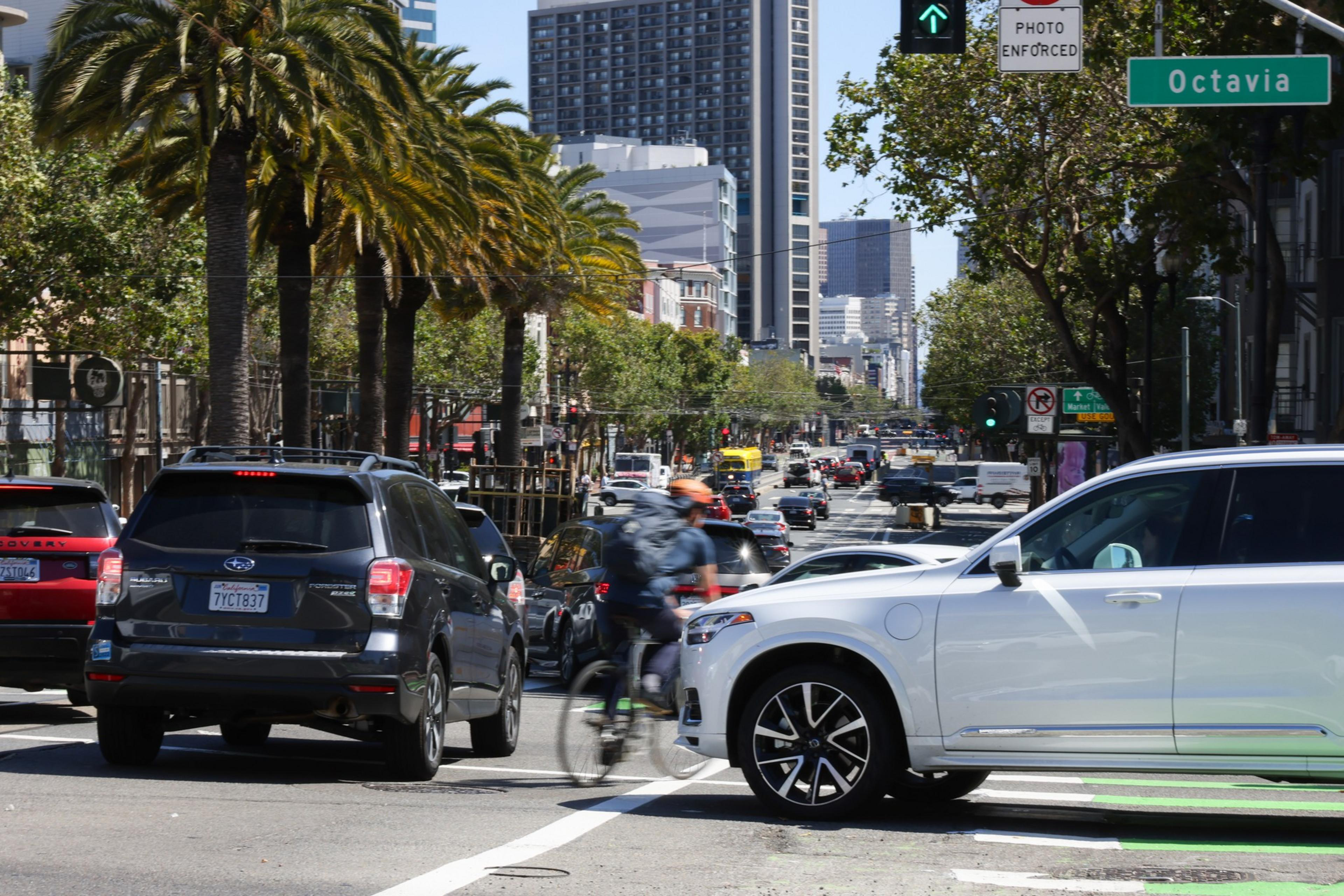 A busy city street with cars stopped at a light, palm trees lining the road, a cyclist wearing a helmet, and a green street sign reading "Octavia."