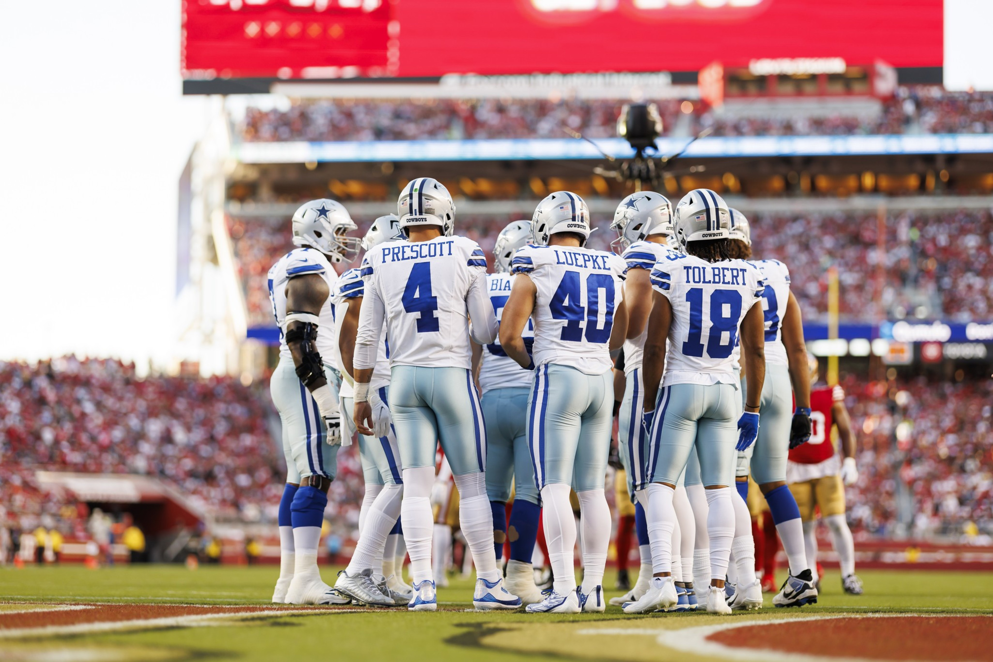 A group of American football players in blue and white uniforms huddle on a grassy field, with a large stadium and cheering crowd in the background.