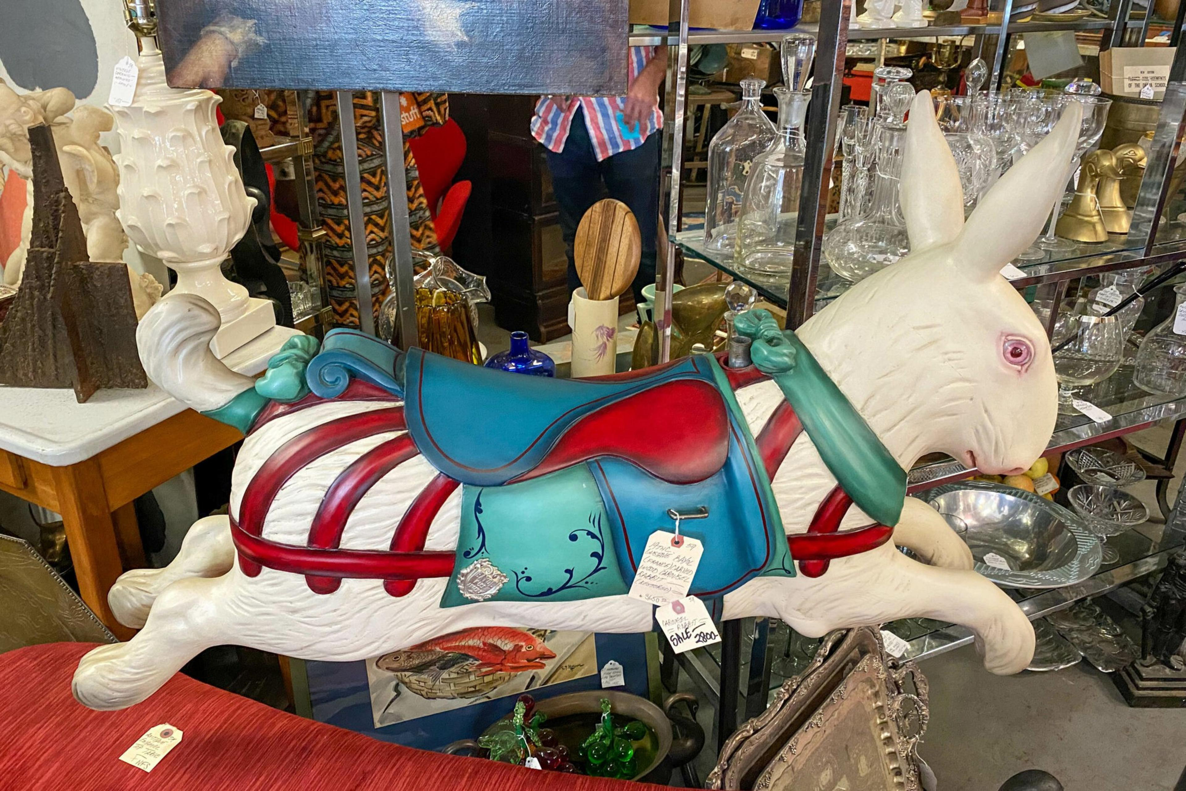 A white, rabbit-shaped carousel figure with a red and blue saddle and green harness sits among various antique items.