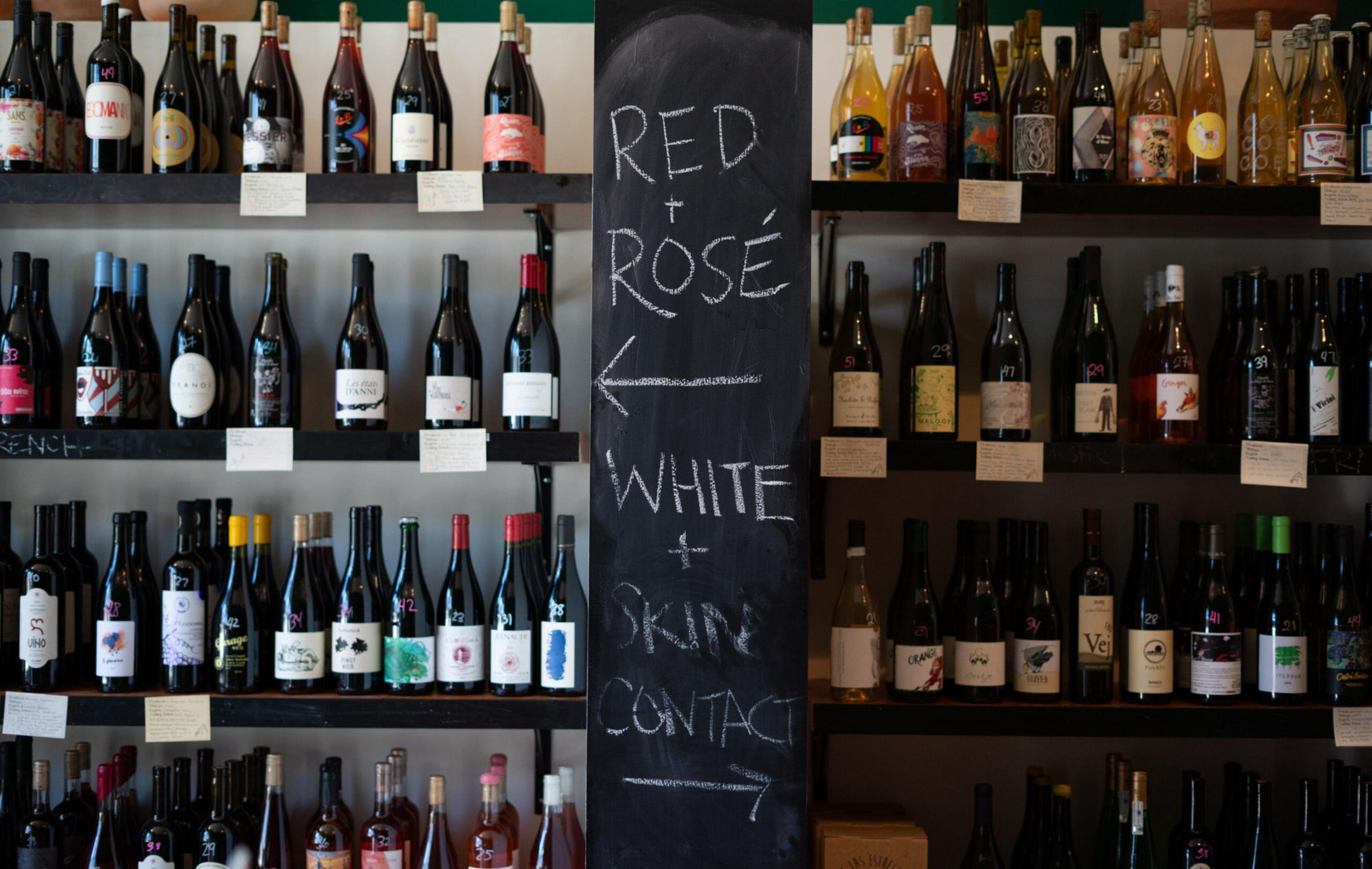 The image shows shelves of wine bottles organized by type. A chalkboard in the center directs "Red + Rosé" to the left and "White + Skin Contact" to the right.