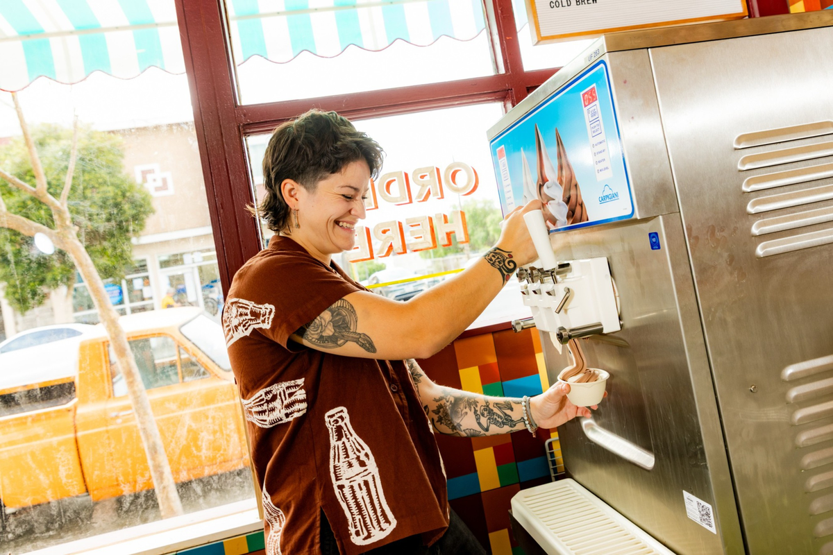 A person with tattoos on their arms smiles while dispensing soft-serve ice cream into a cup from a machine inside a colorful shop.