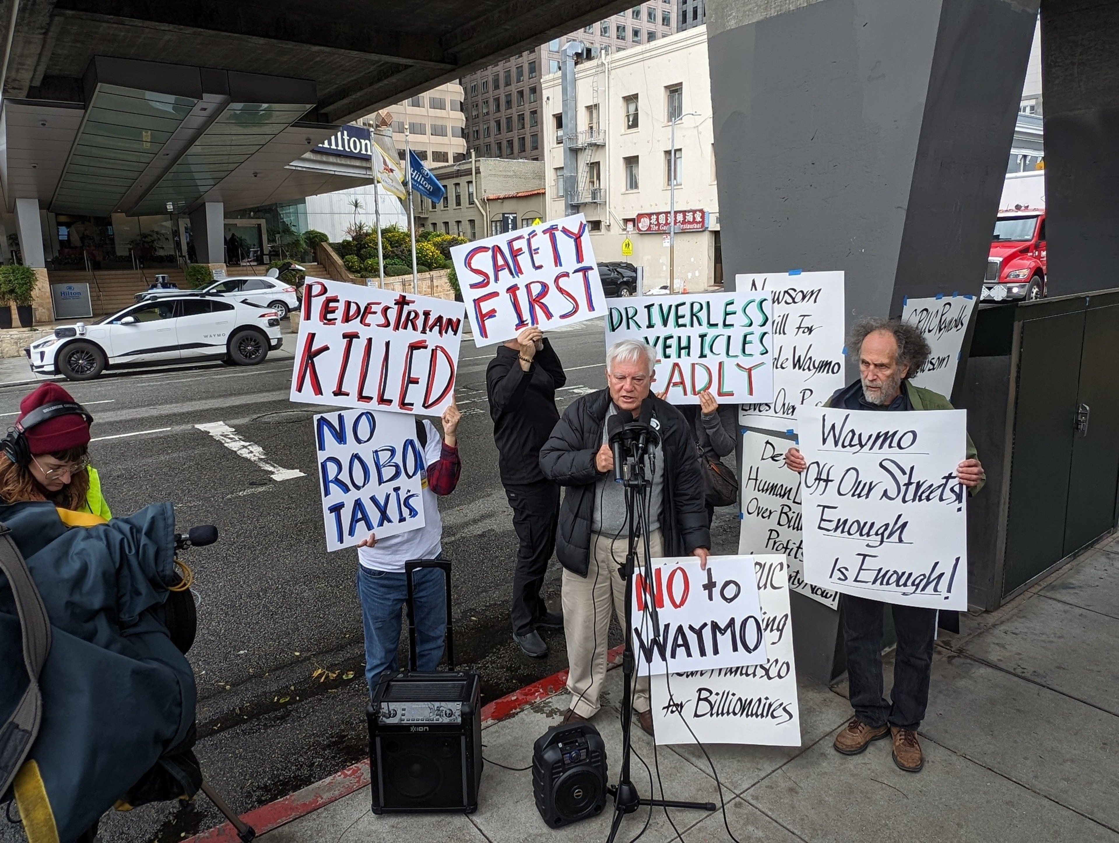 An autonomous vehicle waits outside a hotel as a man holds a posterboard sign and stands next to another speaking into a stand for multiple microphones.